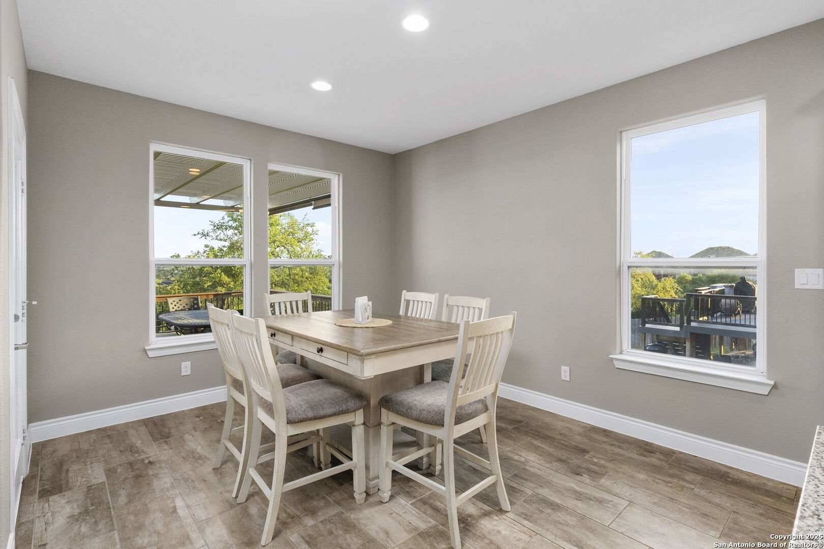 Dining room with a light wood table and chairs, large windows, and views of the deck and trees.