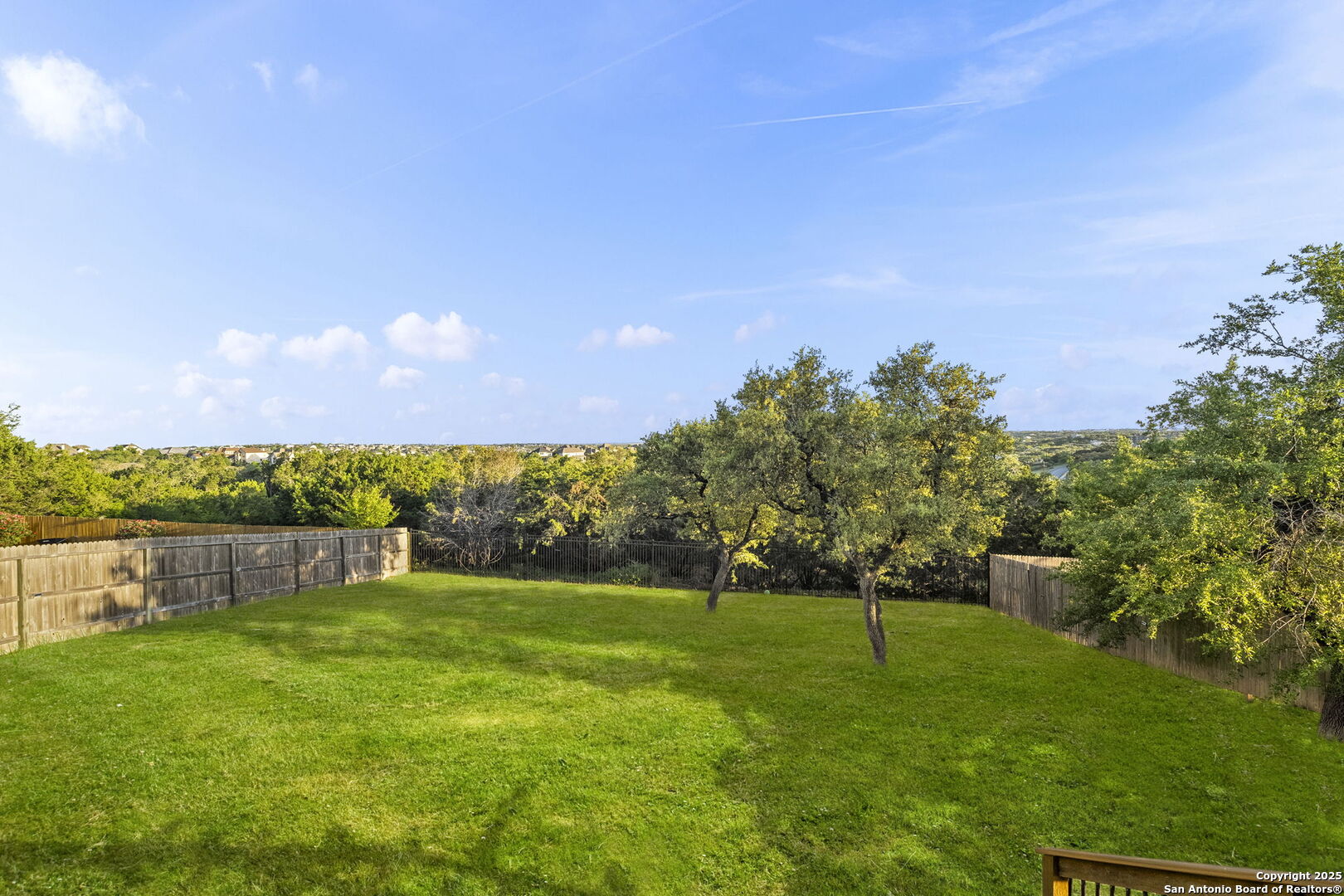 Fenced backyard with a broad green lawn, mature trees, and distant homes across the horizon.