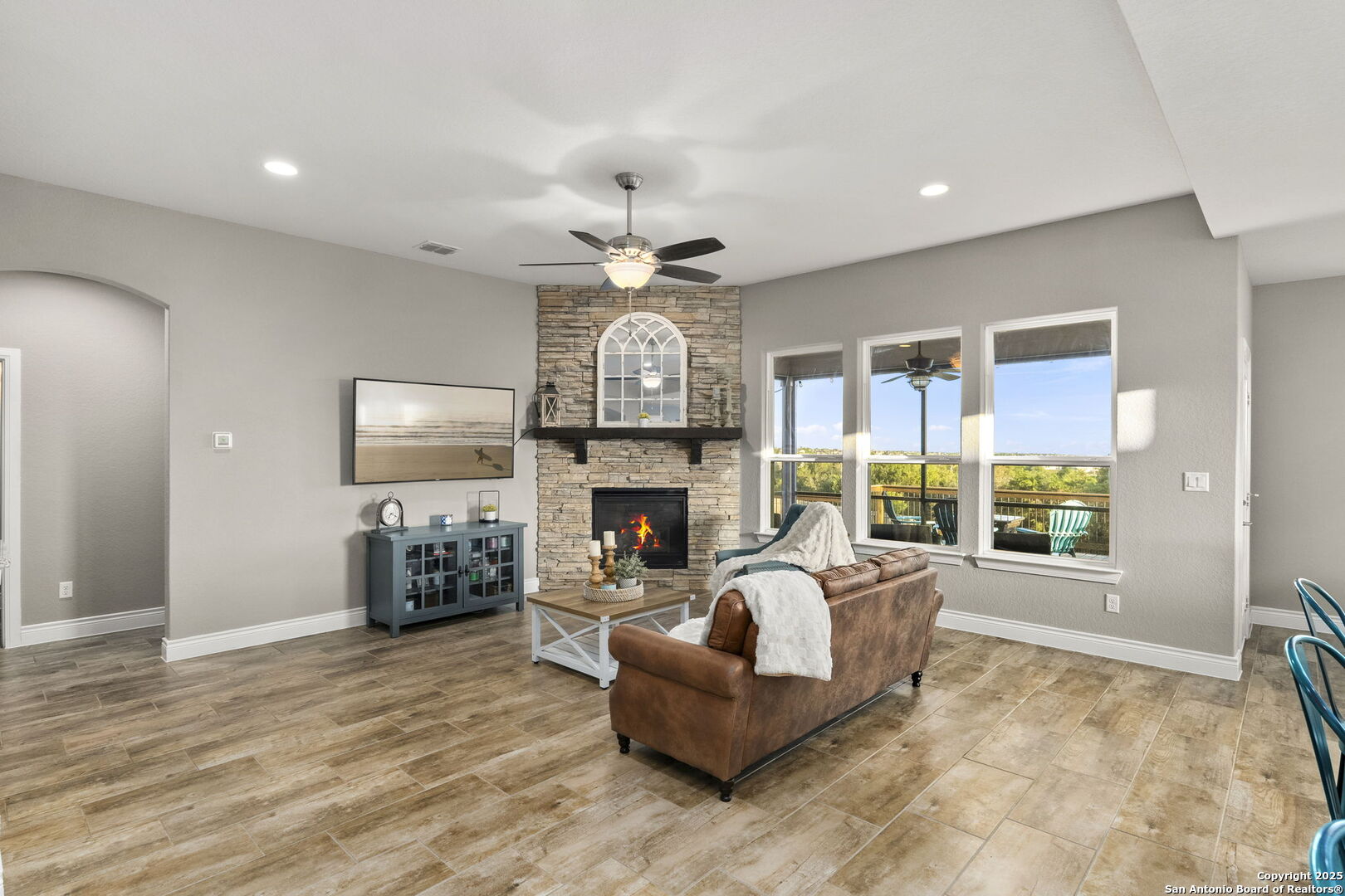 Living room with a stone fireplace, brown tile floors, and large windows overlooking a deck and trees.