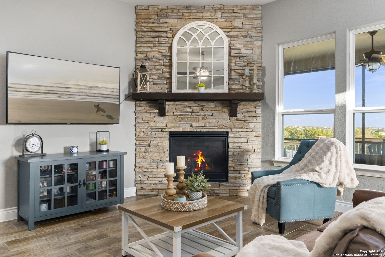 Living room with a stacked-stone fireplace, teal armchair, and large windows overlooking greenery.