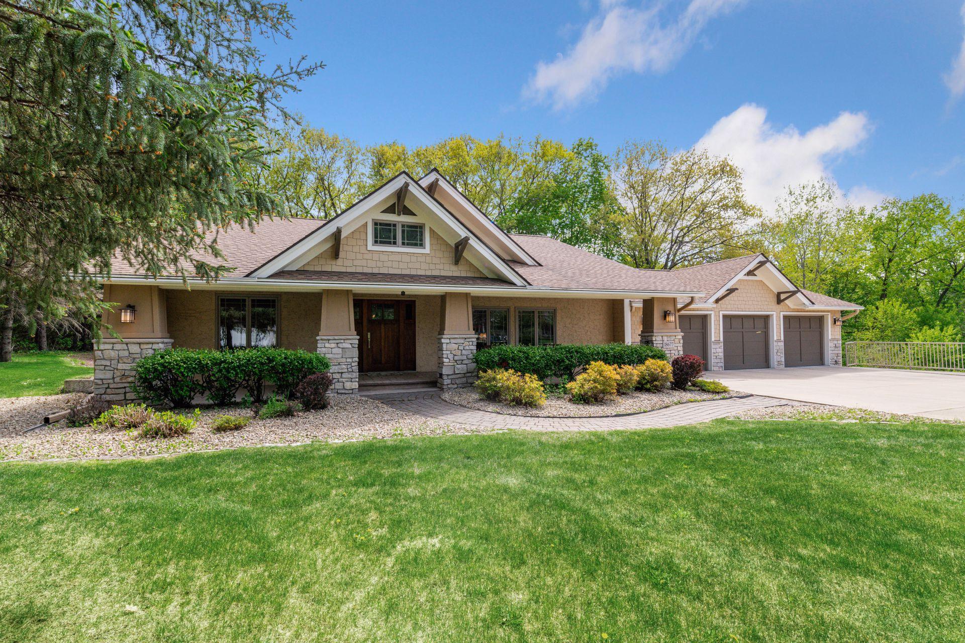 Beige single-story home with stone porch columns, a three-car garage, and a broad green lawn.