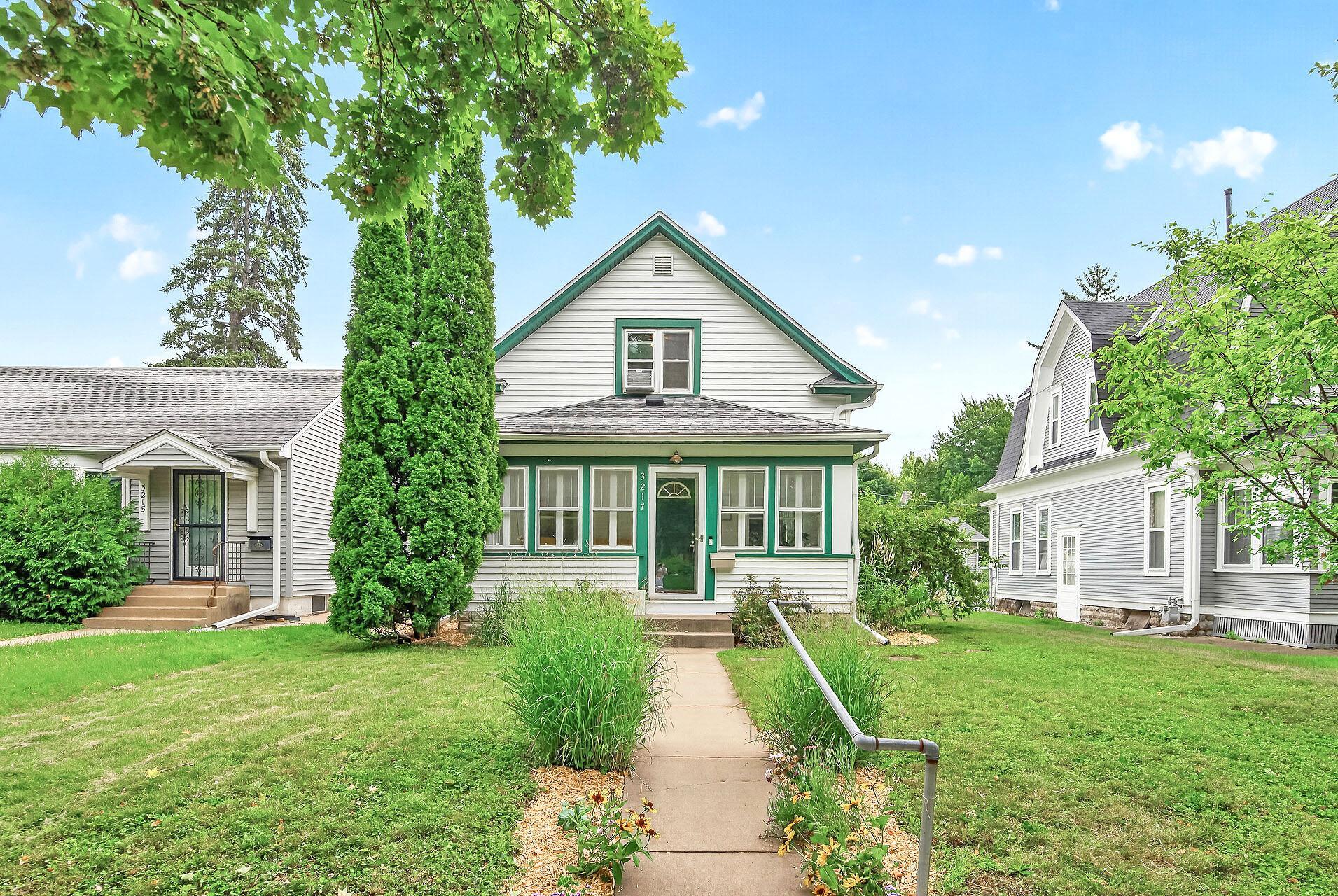 White gable-front house with green trim, a central walkway, and mature trees.