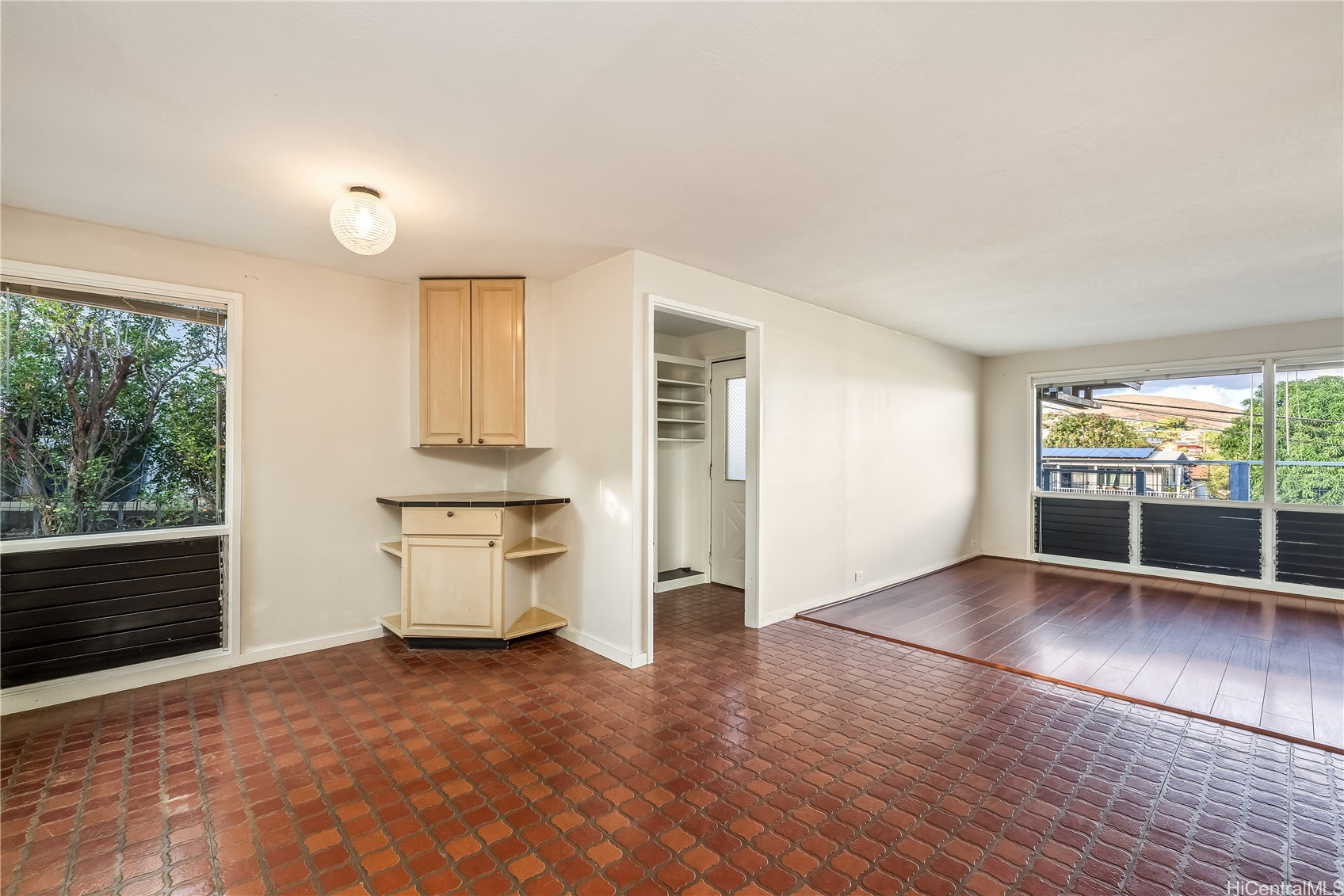 Vacant room with brick-pattern tile flooring, corner cabinet, and windows beside an adjoining wood-floored area.