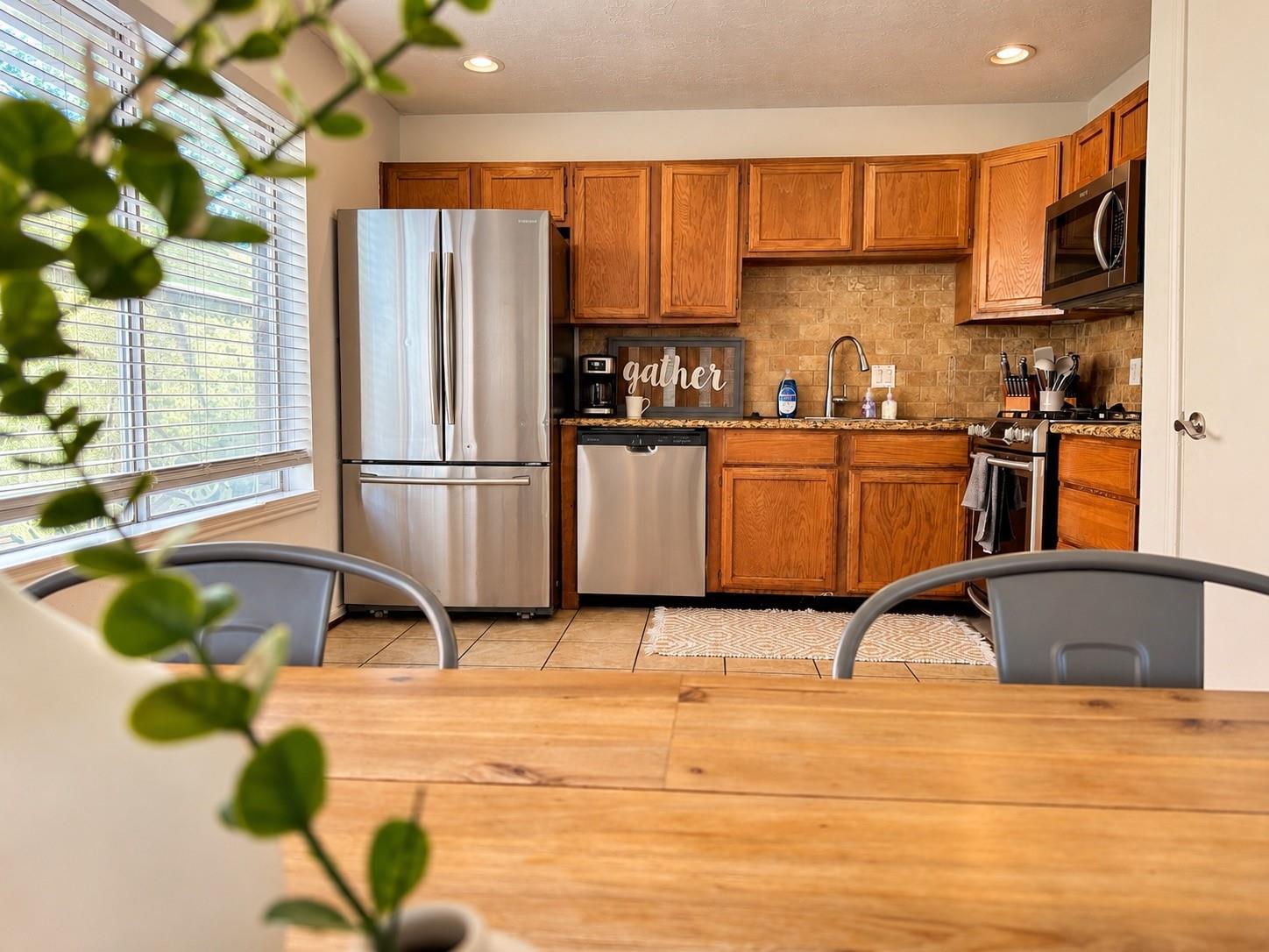 Kitchen with warm wood cabinets, stone backsplash, stainless steel appliances, and a window with blinds.