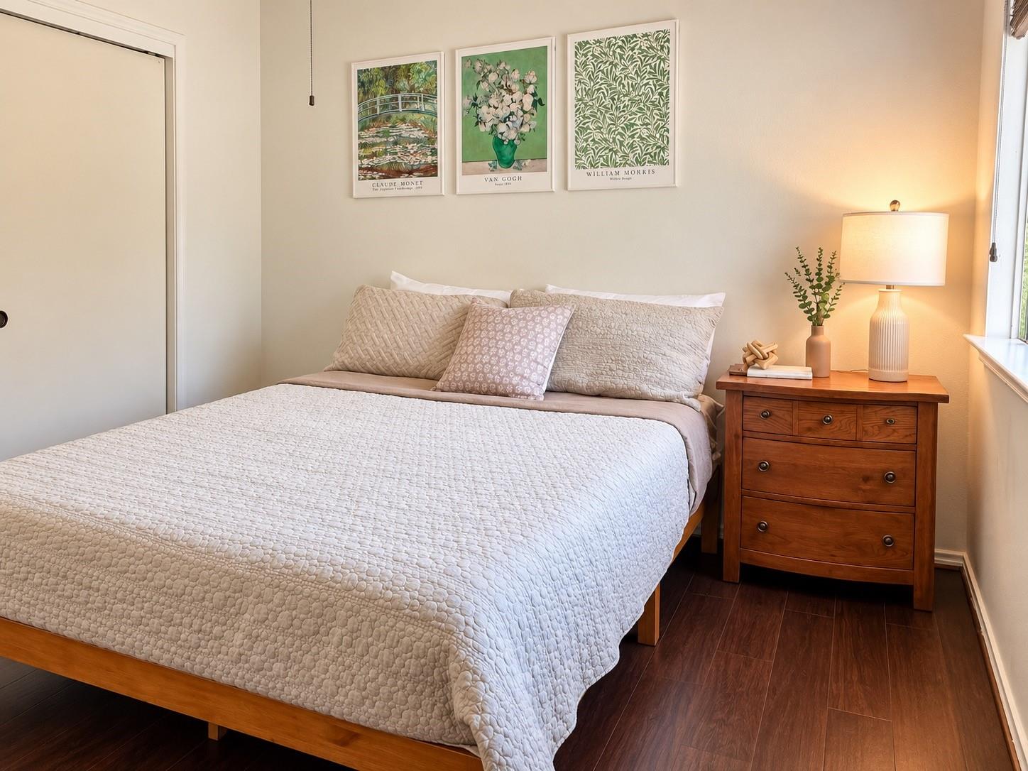 Bedroom with a white quilted bed, three framed prints, and a wooden bedside chest with a lamp.