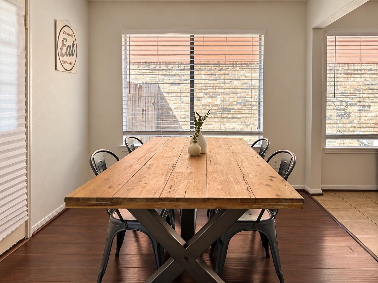 Dining room with a long rustic wood table, metal chairs, and wide windows with blinds.