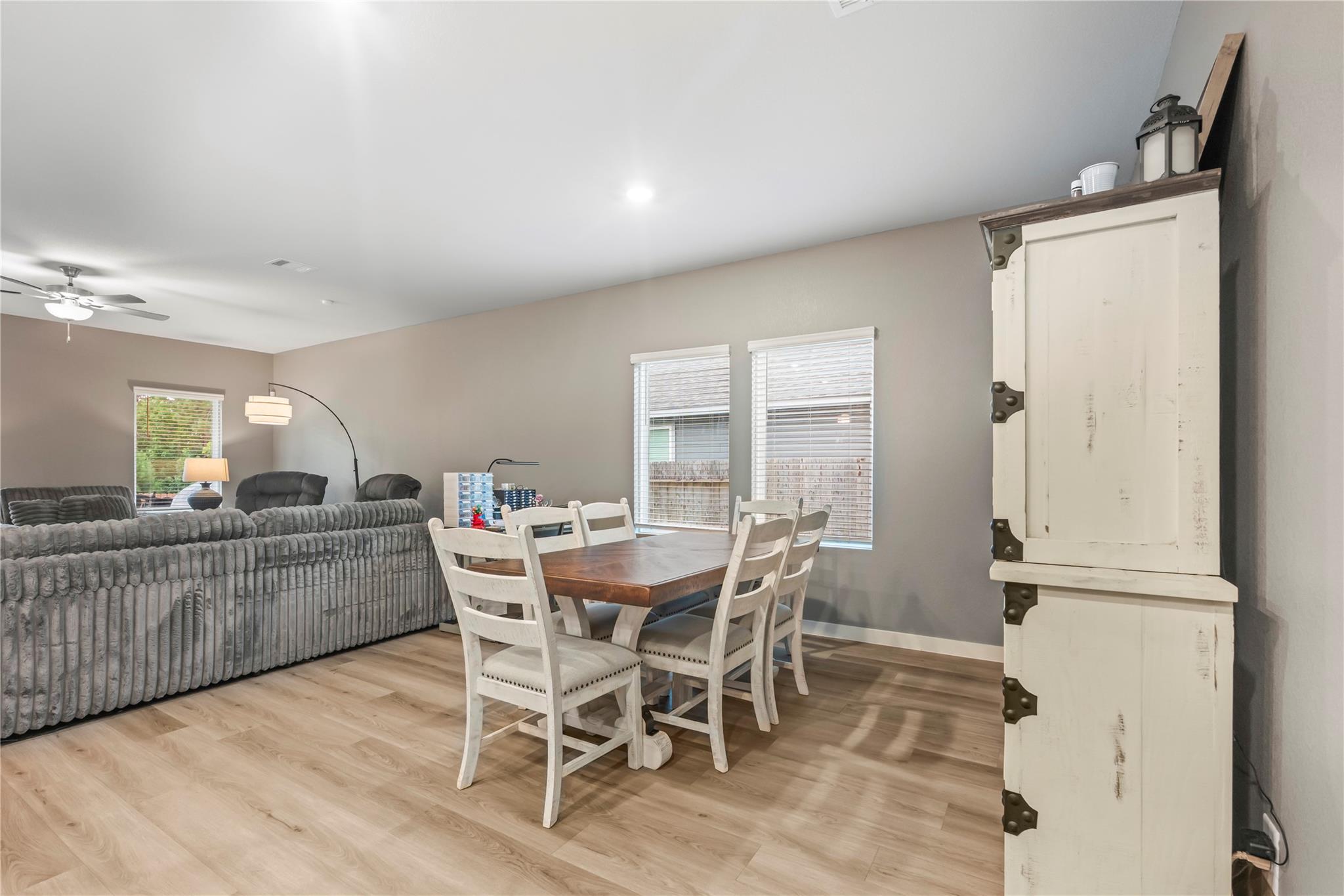 Open-plan living and dining area with a wooden table, white chairs, gray sofas, and a distressed white cabinet.