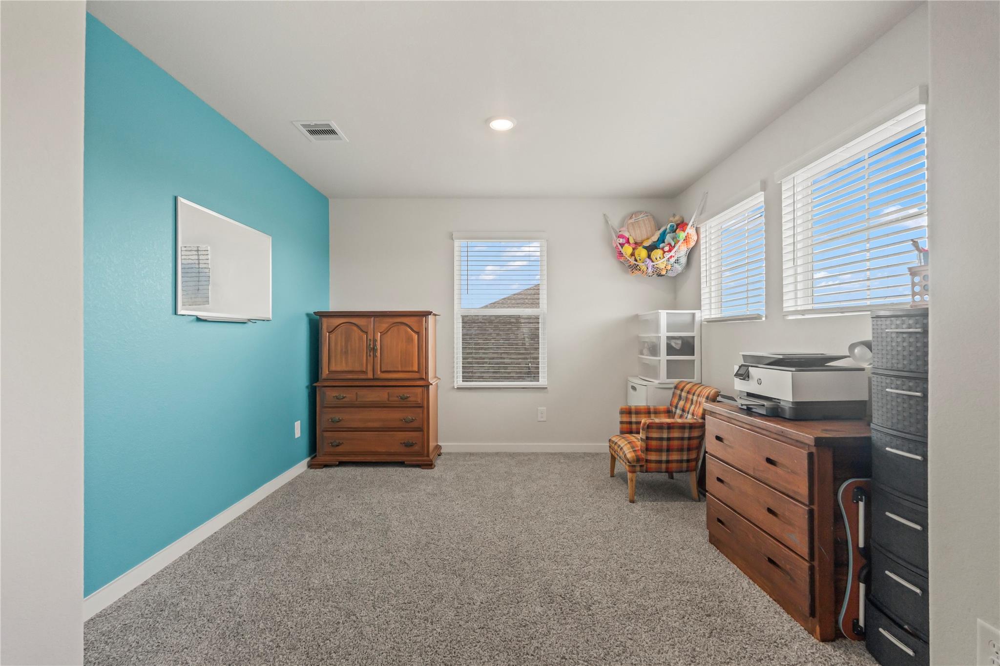 Carpeted bedroom with a turquoise accent wall, wooden dressers, plaid chair, and windows with blinds.