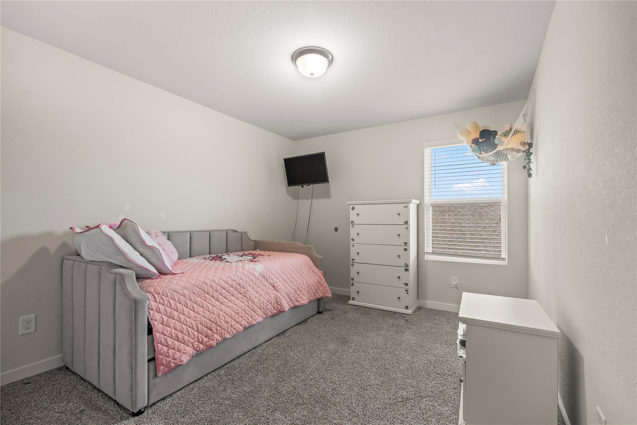 Furnished bedroom with a gray bed, pink quilt, white dresser, and wall-mounted television.