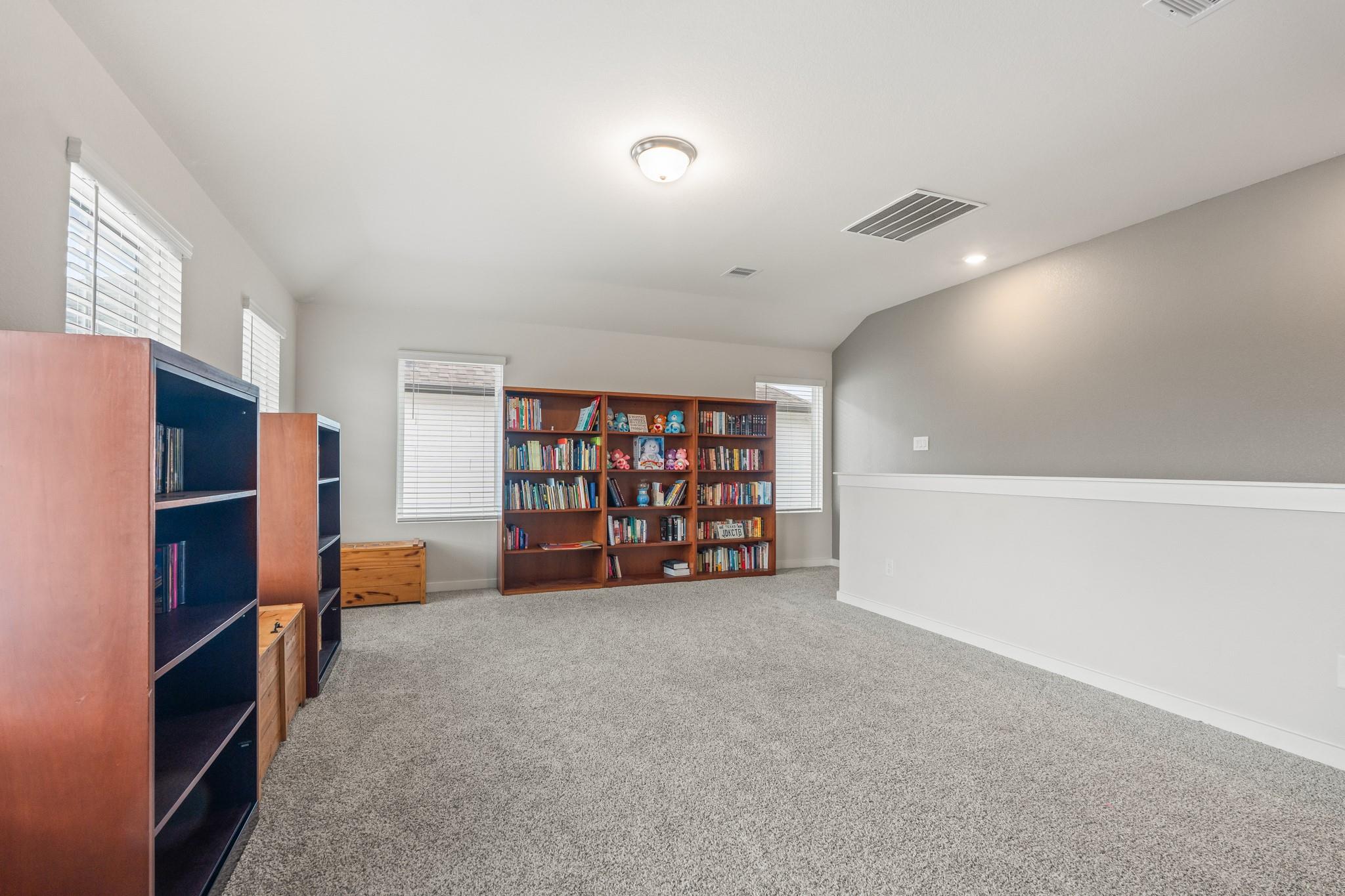 Carpeted bonus room with book-filled shelves, toys, and windows with blinds.