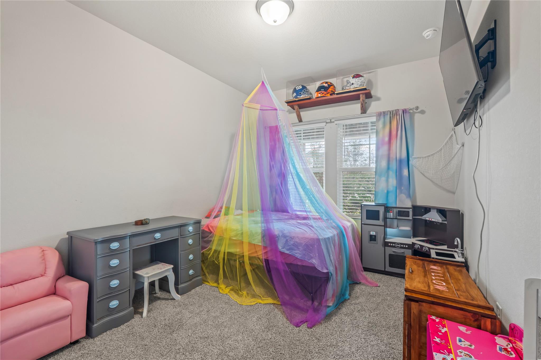 Child's bedroom with a rainbow canopy bed, gray desk, pink armchair and toy kitchen.
