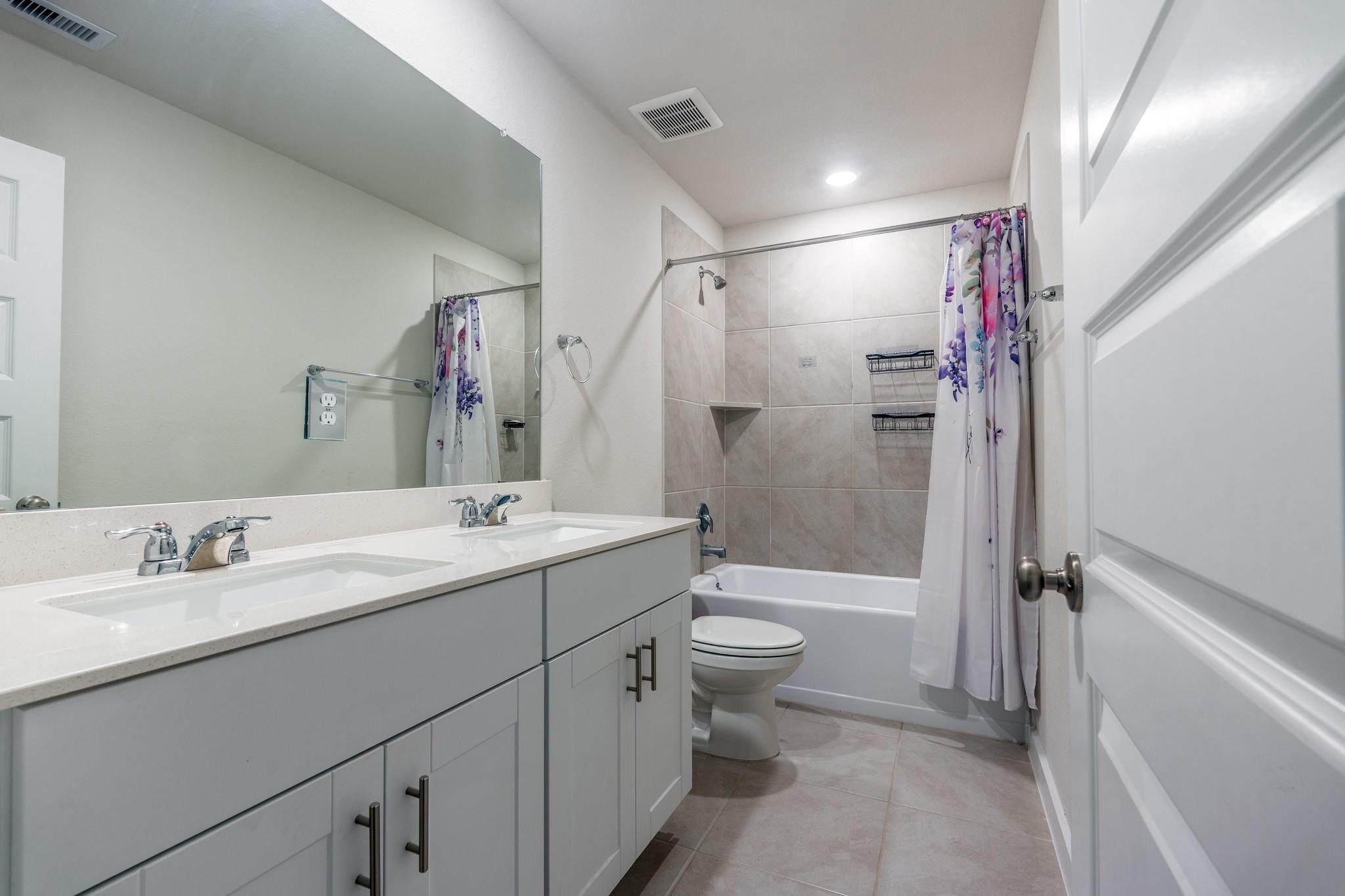 Bathroom with a double-sink vanity and tiled tub-shower behind a floral curtain.