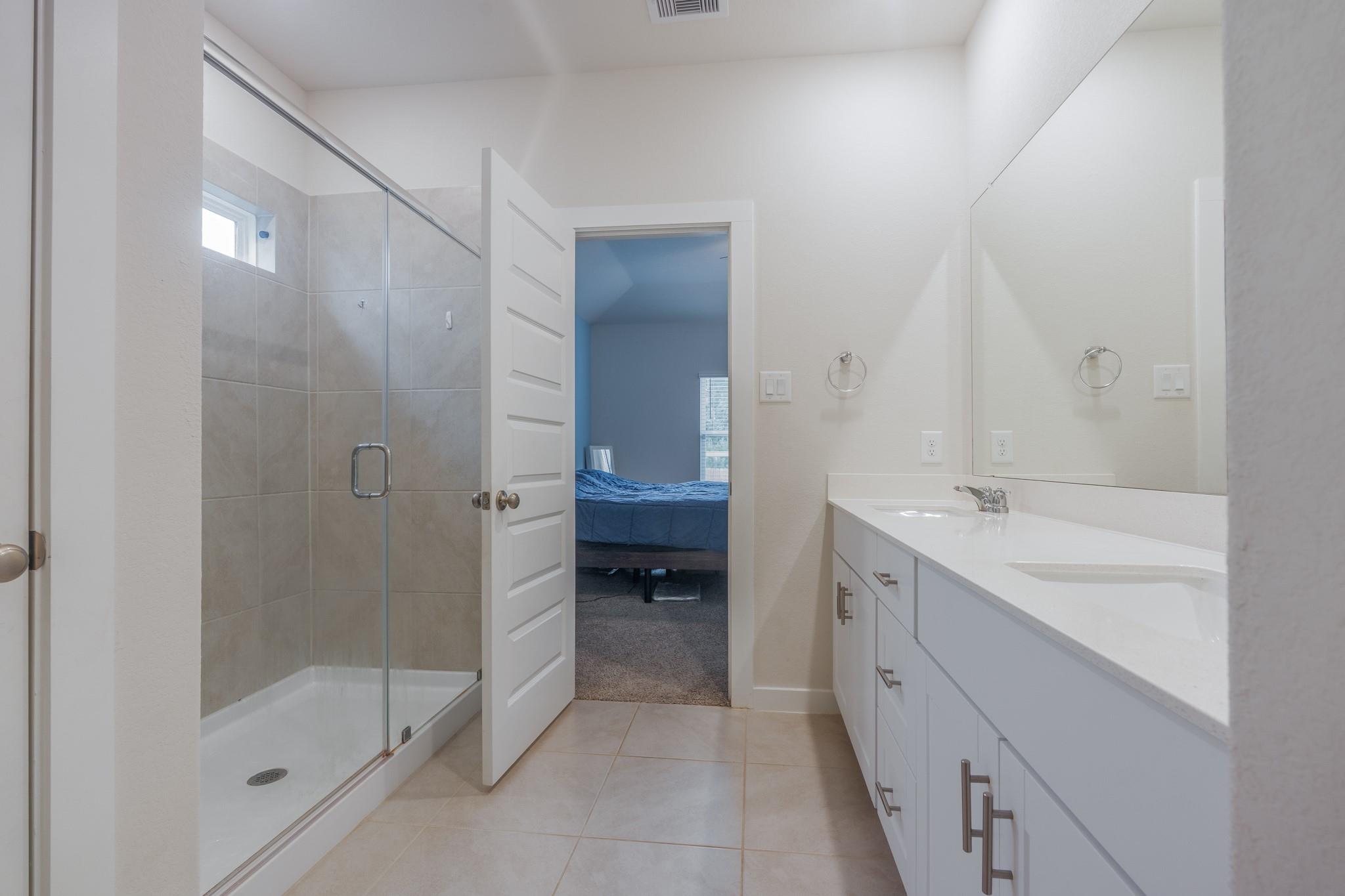 Bathroom with a glass-enclosed shower and long double-sink vanity, opening onto a bedroom.