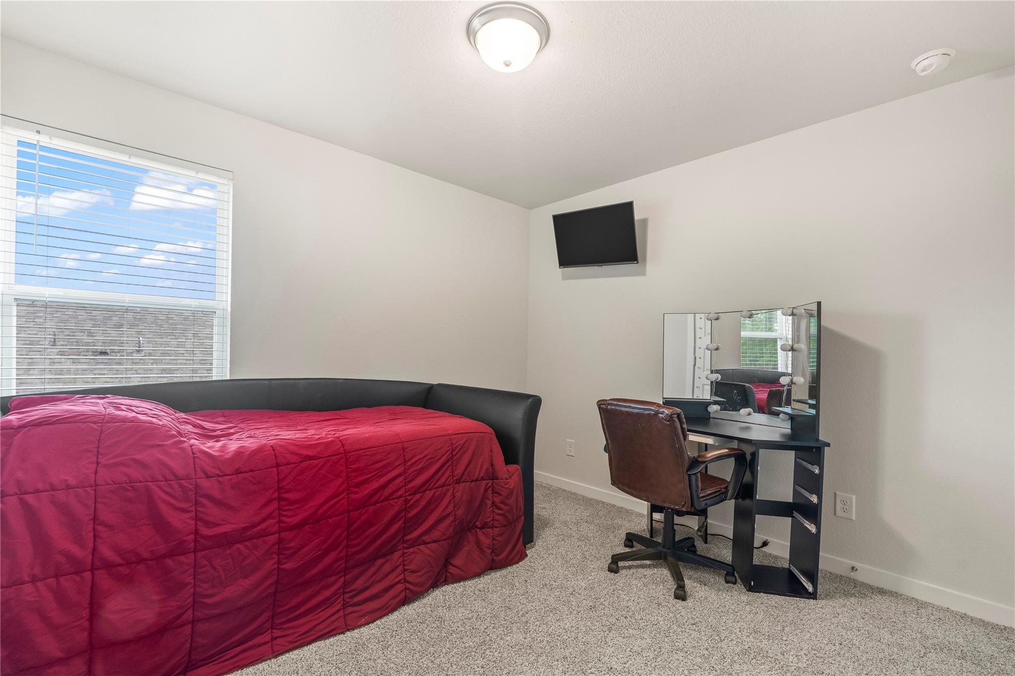 Bedroom with a magenta-covered bed, window with blinds, and vanity desk with lighted mirror.