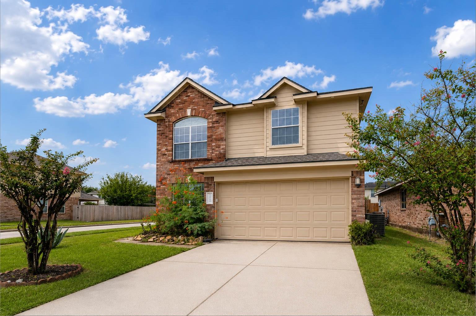 Two-story tan-and-brick house with a two-car garage, broad driveway, and flowering shrubs.