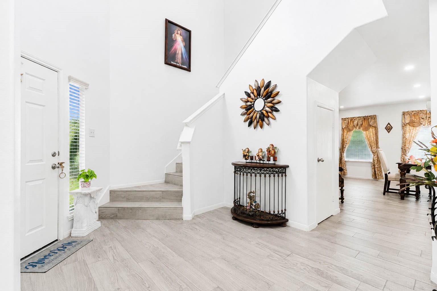 Bright foyer with a white staircase, pale tile floors, and a decorative console beneath a sunburst mirror.
