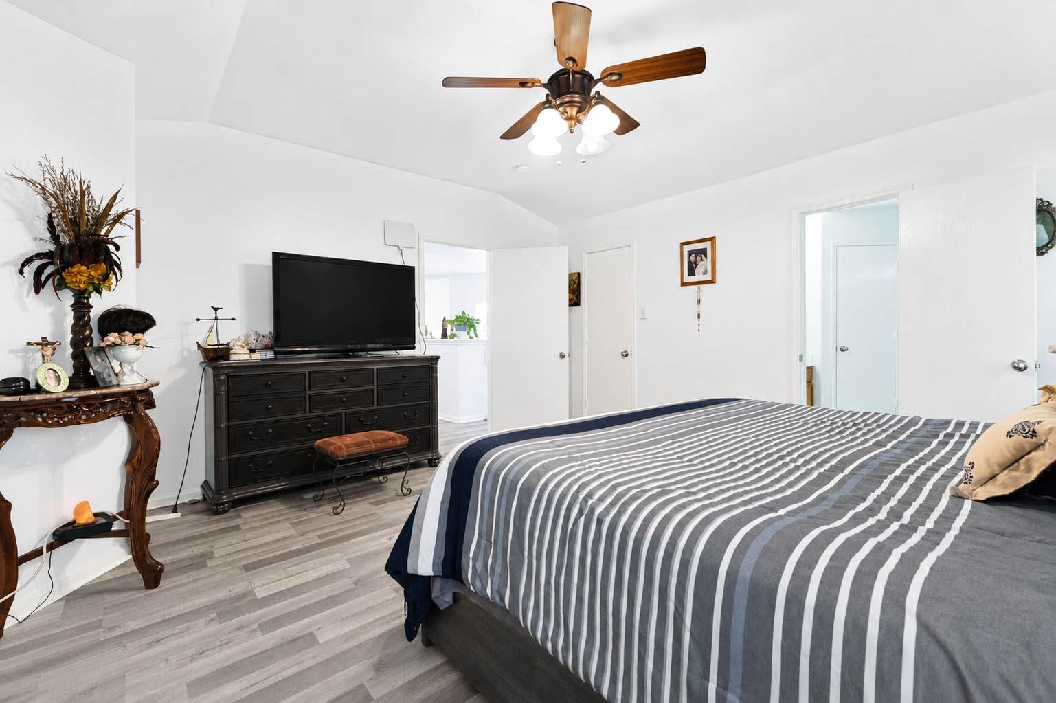 Furnished bedroom with a gray-and-white striped bedspread, dark dresser and TV, and wood-bladed ceiling fan.