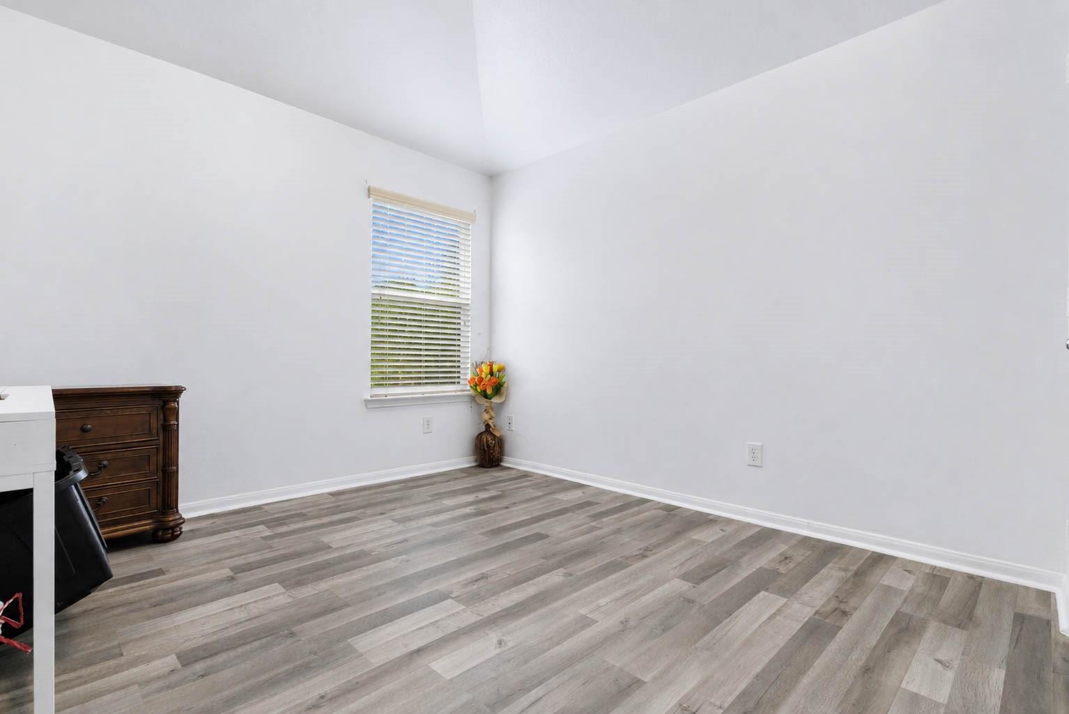 Mostly empty room with gray plank flooring, white walls, and a window with blinds.