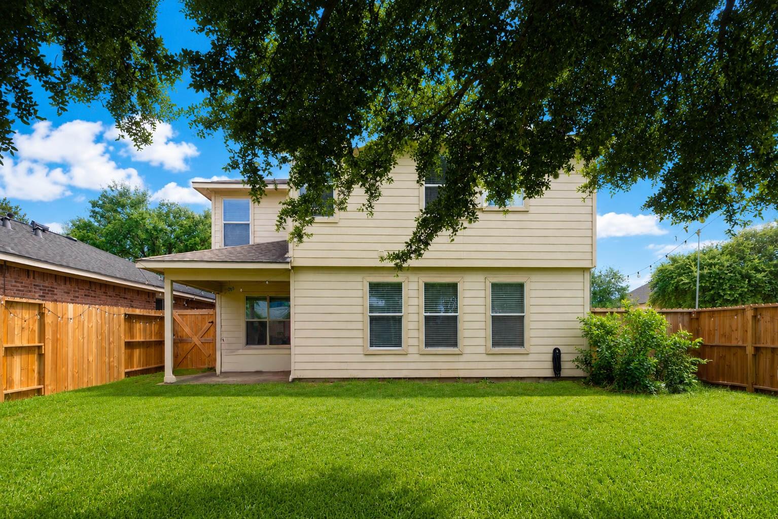 Rear of a two-story beige house behind a fenced lawn, shaded by mature trees.