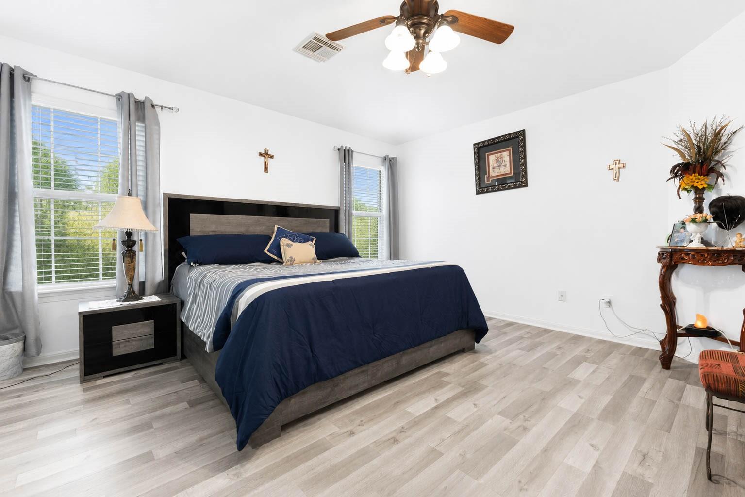 Staged bedroom with a navy-covered bed, gray flooring, two curtained windows, and a ceiling fan.