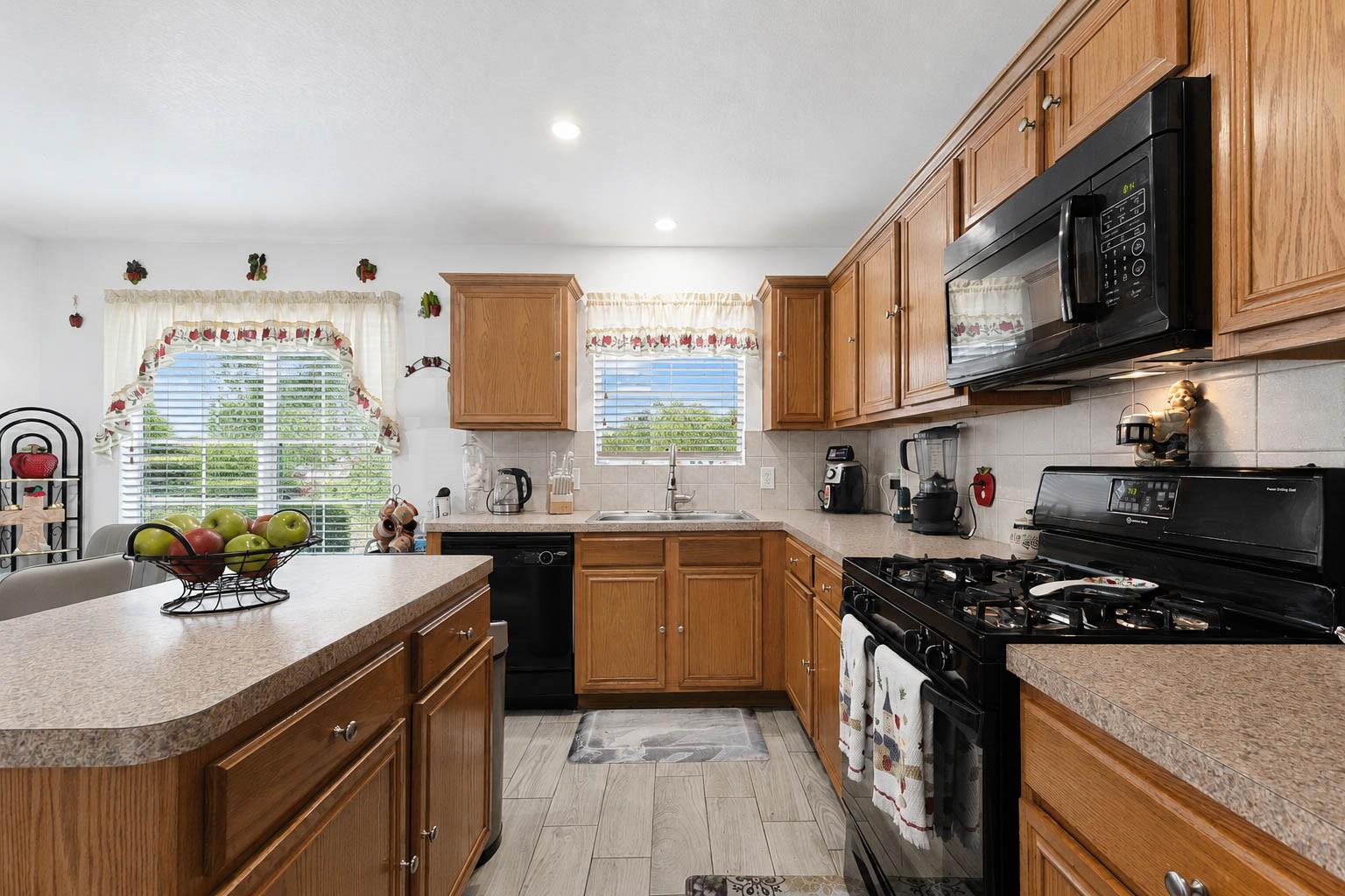 Kitchen with honey-toned wood cabinets, black appliances, and a fruit bowl on the foreground counter.