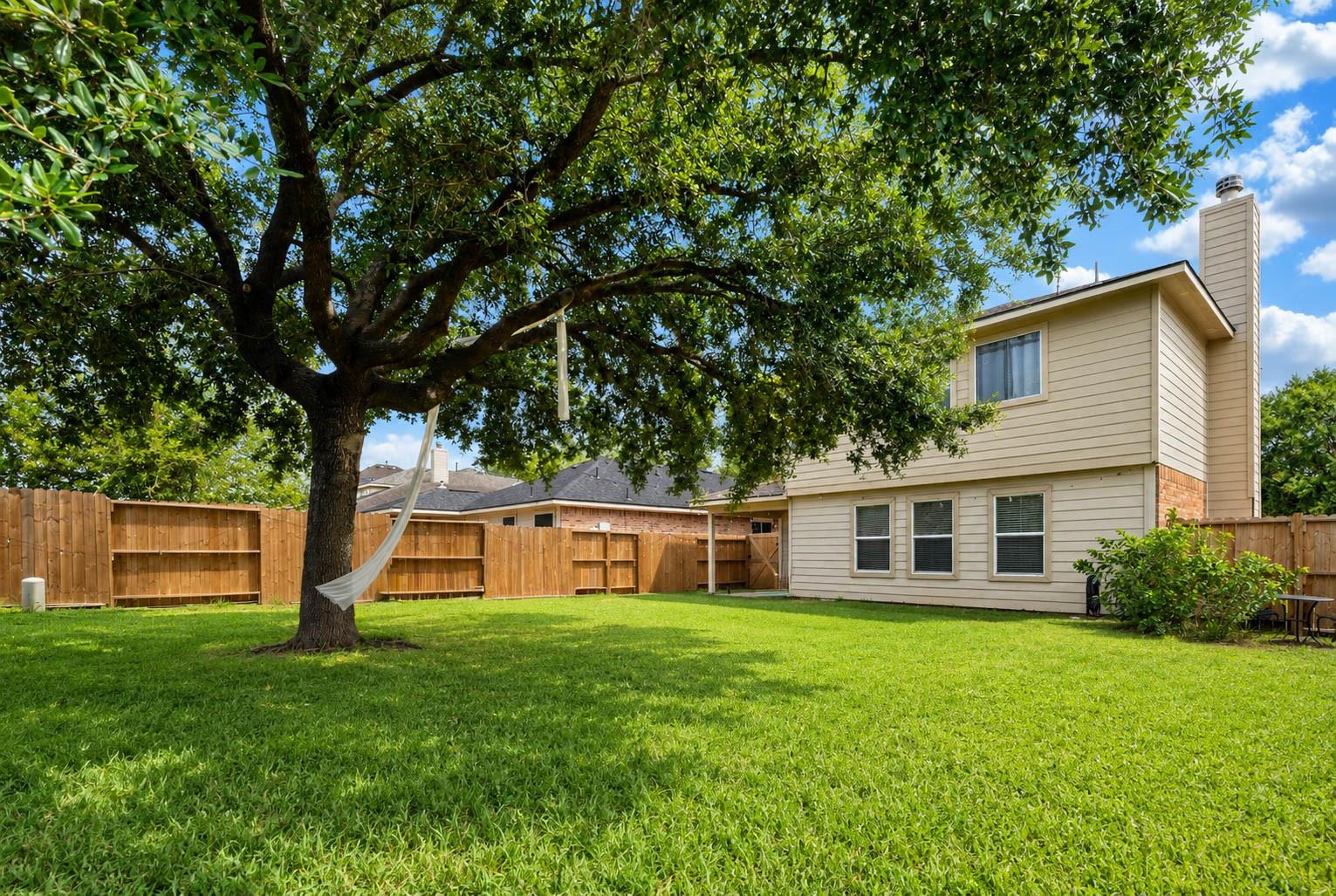 Two-story home behind a fenced lawn, shaded by a large tree with a hammock.