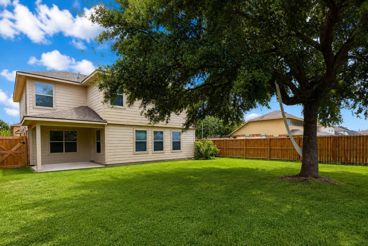 Two-story beige house behind a broad lawn, with a covered patio, wooden fence, and large shade tree.
