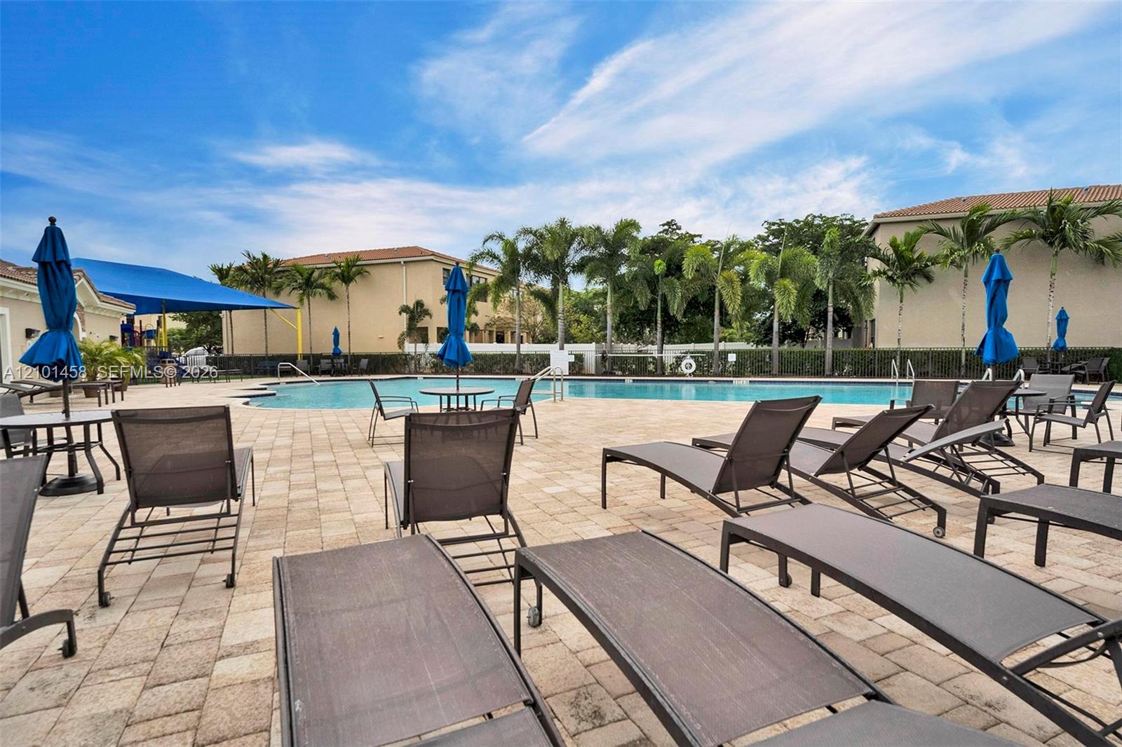 Community pool with a broad paved deck, lounge chairs, blue umbrellas, and palm trees.