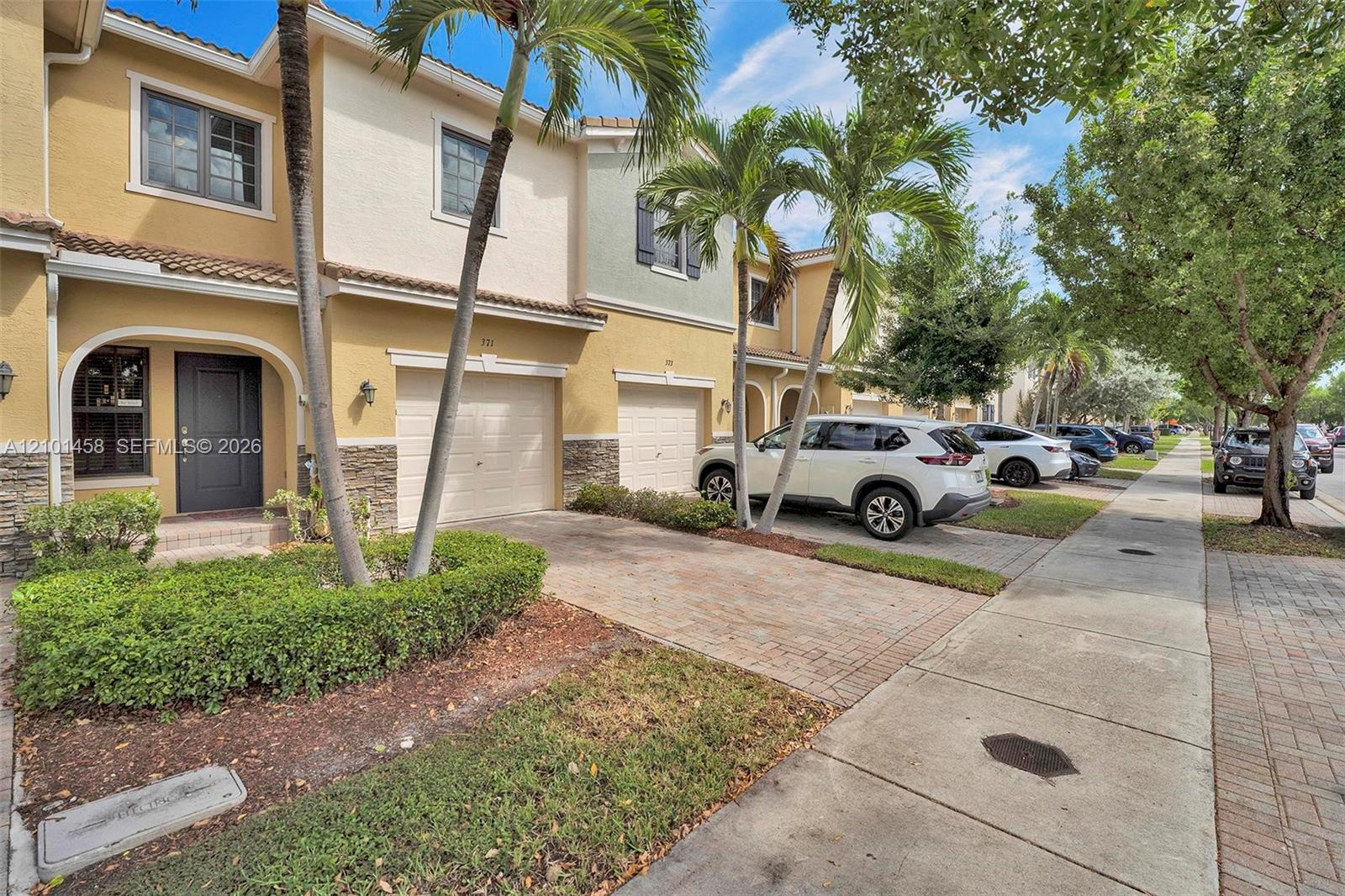 Attached two-story townhomes with garages and palm trees along a sunny sidewalk.
