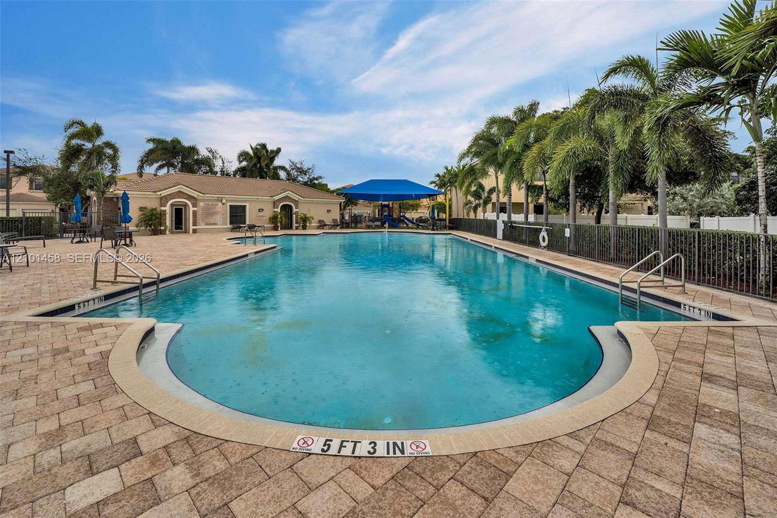 Community pool with a curved shallow end, paver deck, palm trees, and a blue-canopy playground.