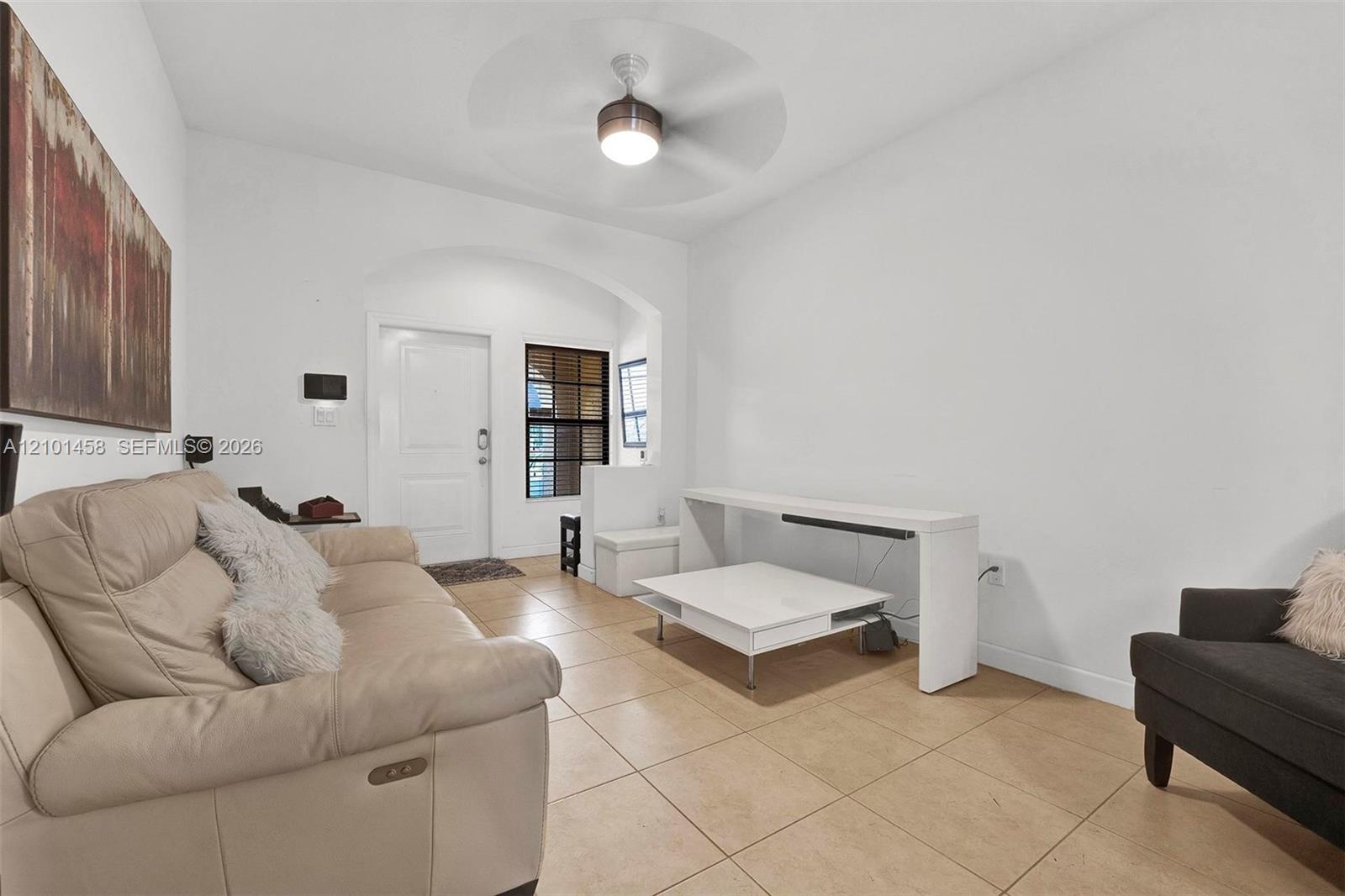 Furnished living room with beige sofa, white tables, tile floors, and a ceiling fan.