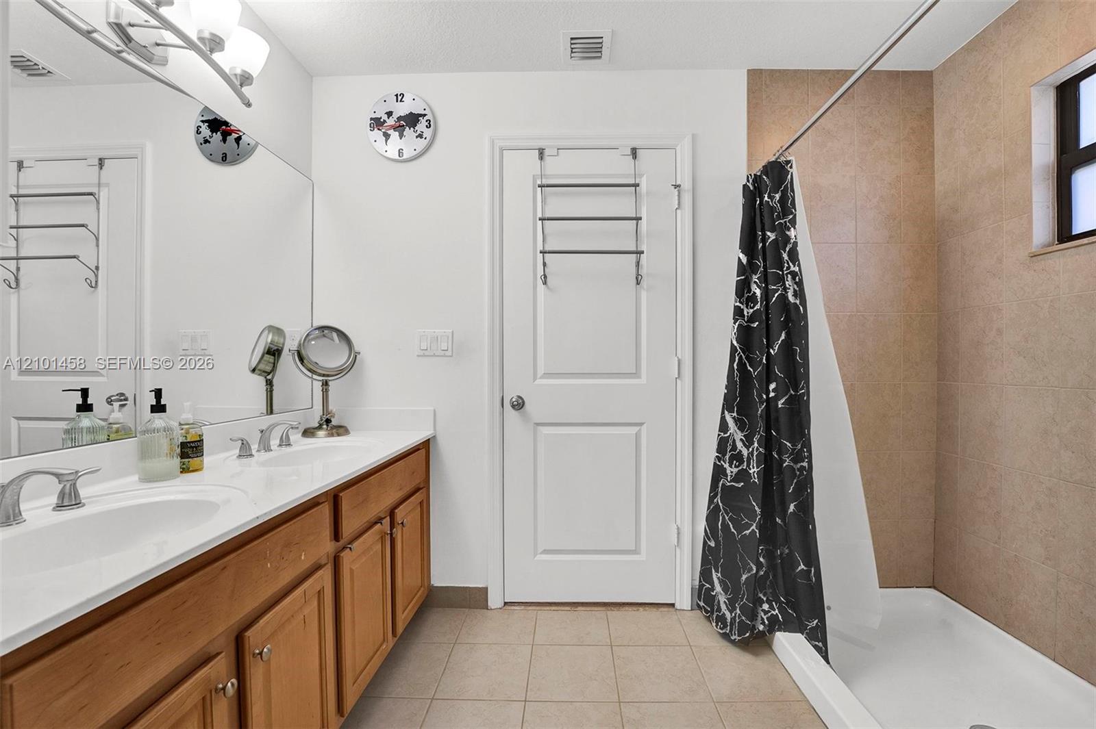 Bathroom with a wood double vanity, tan-tiled tub and shower, and black patterned curtain.