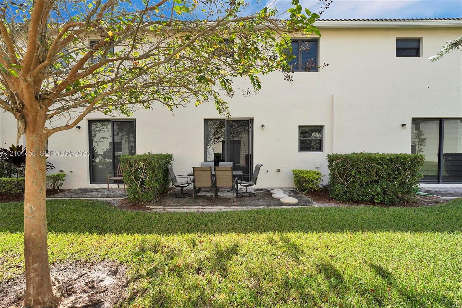 Rear of a two-story white home with a furnished patio, lawn, hedges, and a leafy tree.