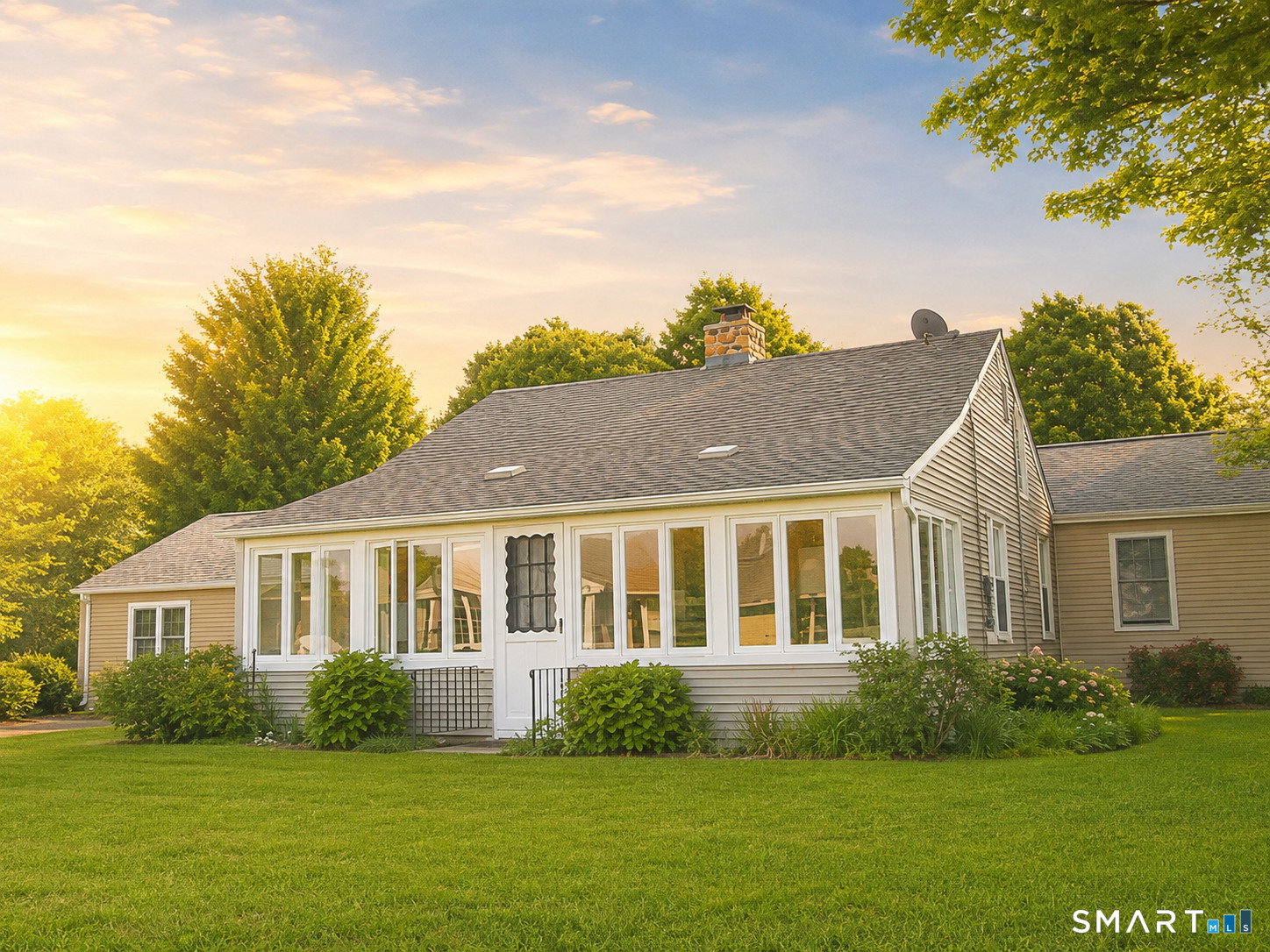 House with a wide, windowed sunroom, stone chimney, and green lawn in golden-hour light.