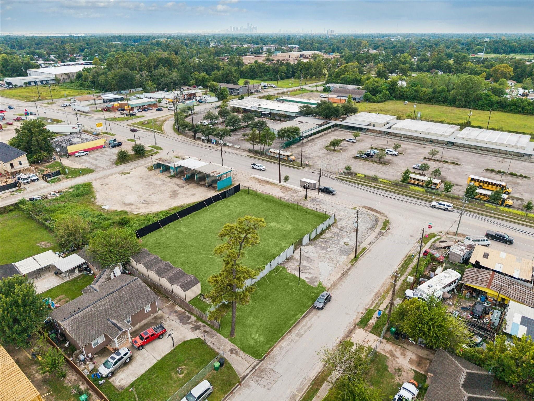 Aerial view of a fenced grassy lot at a road intersection, with commercial buildings and the distant Houston skyline.
