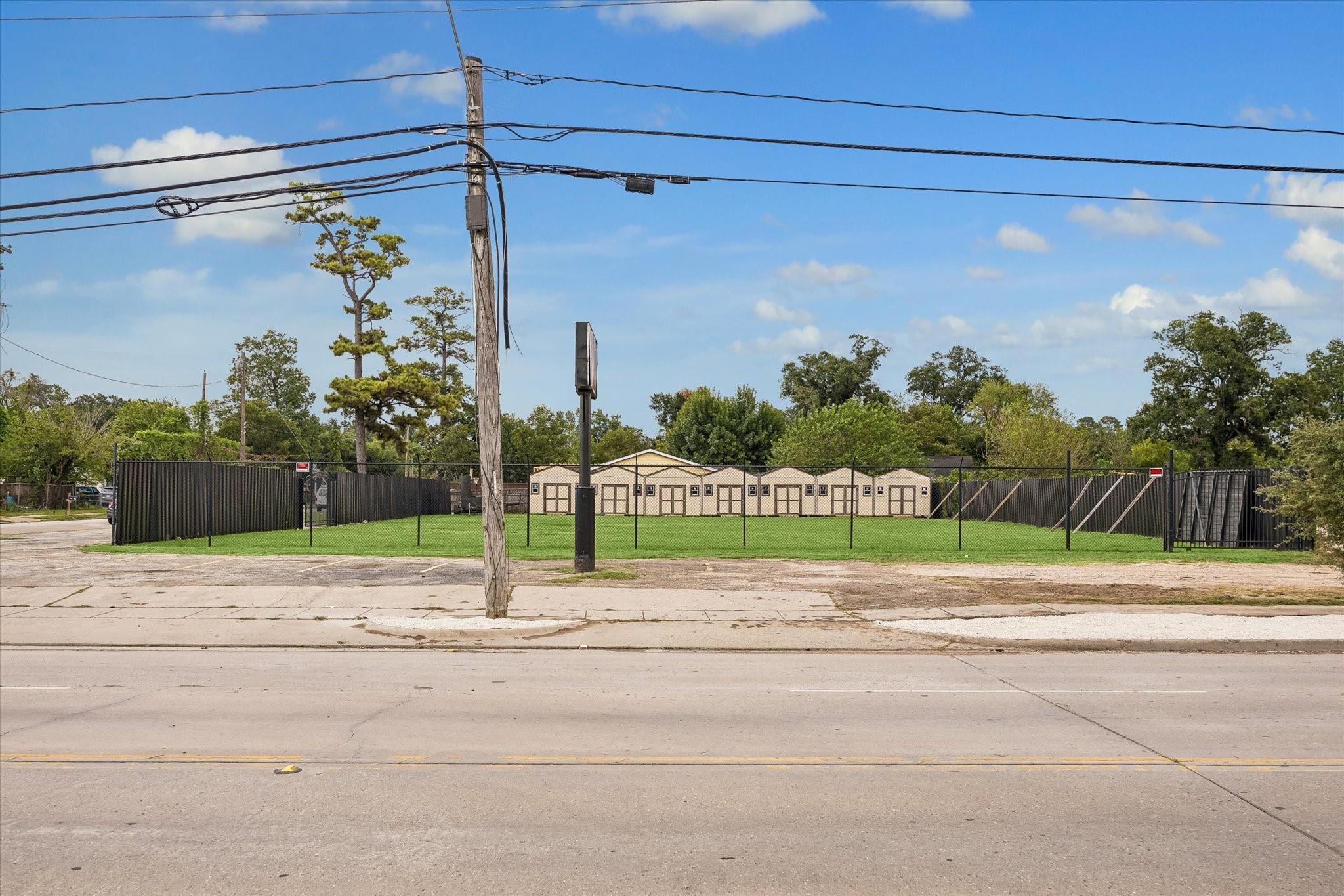 Fenced property with a row of beige buildings behind a grassy yard and roadside utility wires.