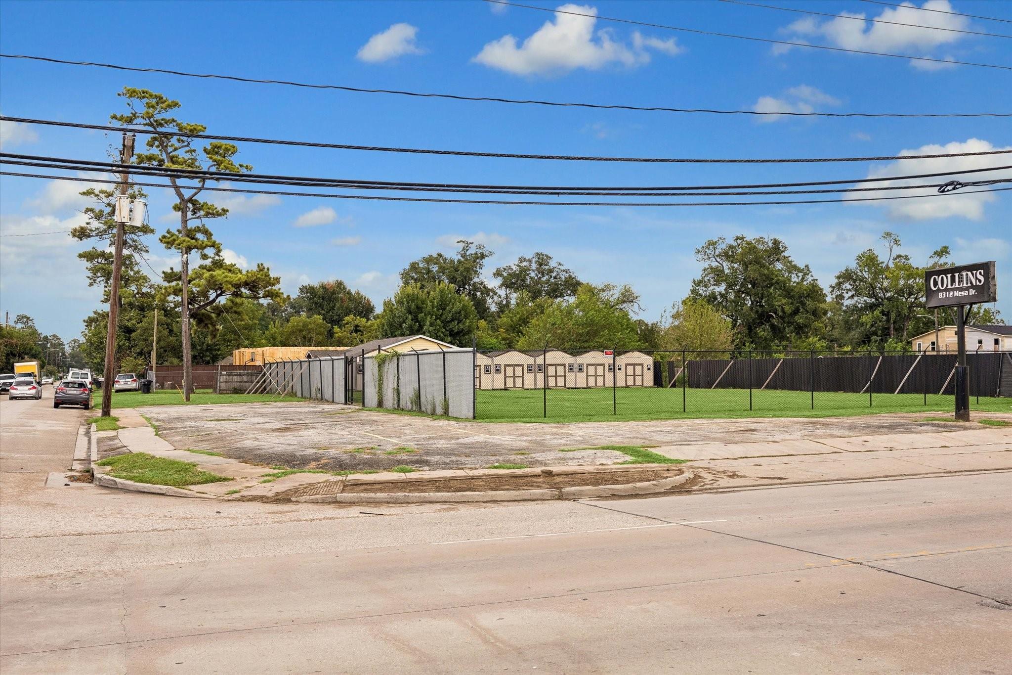 Fenced commercial property with a grassy yard, low buildings, and paved parking area beside the street.