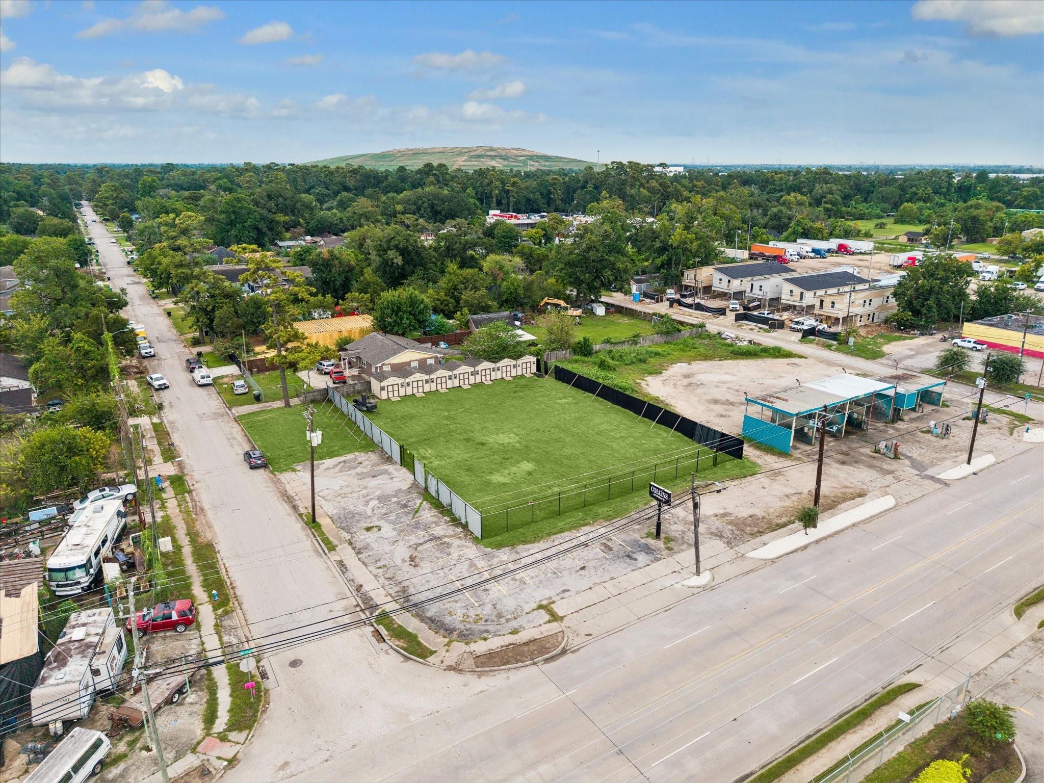 Aerial view of a fenced grassy lot at a street corner, with paved frontage and nearby buildings.
