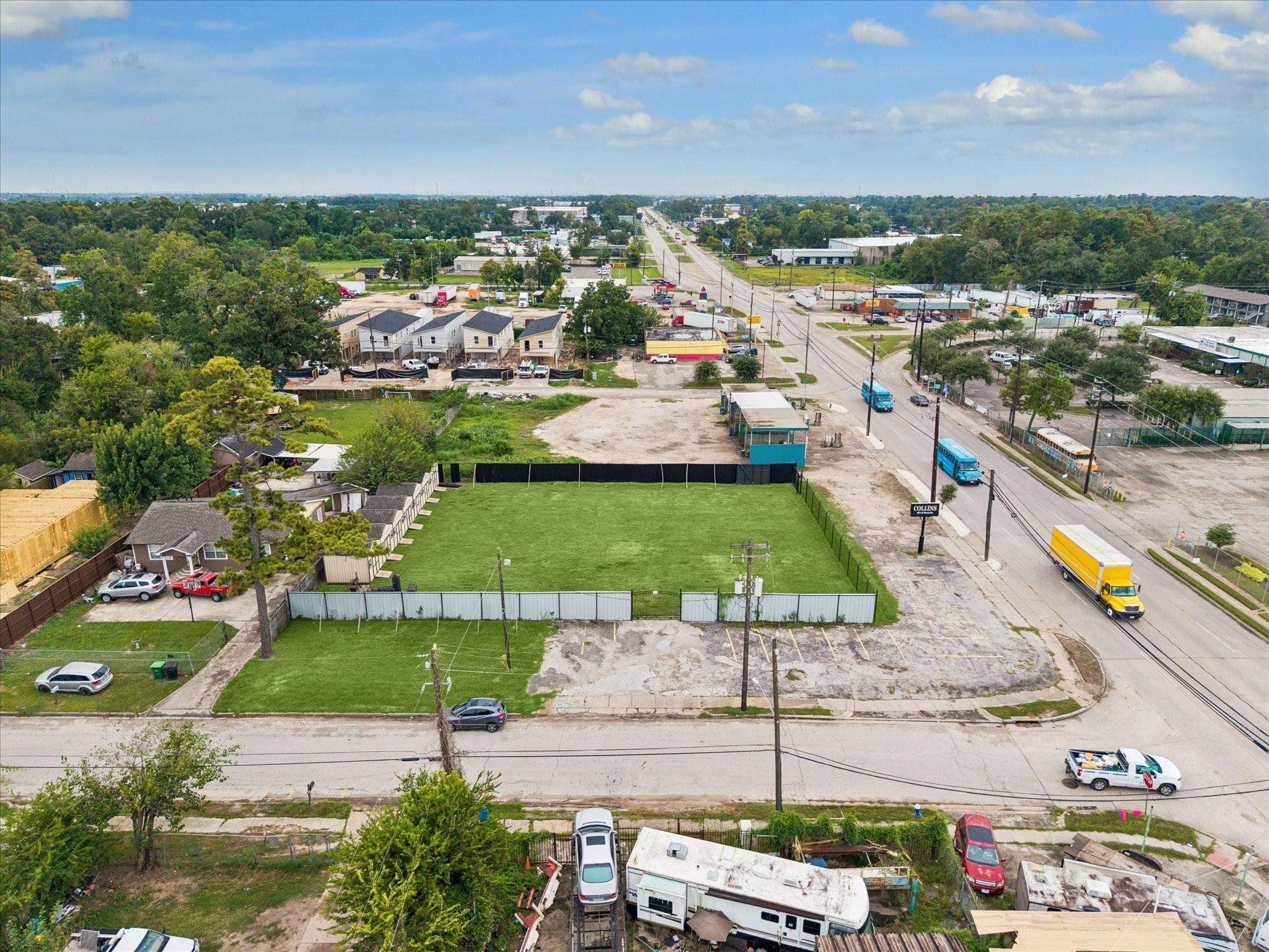 Fenced grassy vacant lot at a road intersection, with nearby homes and businesses.