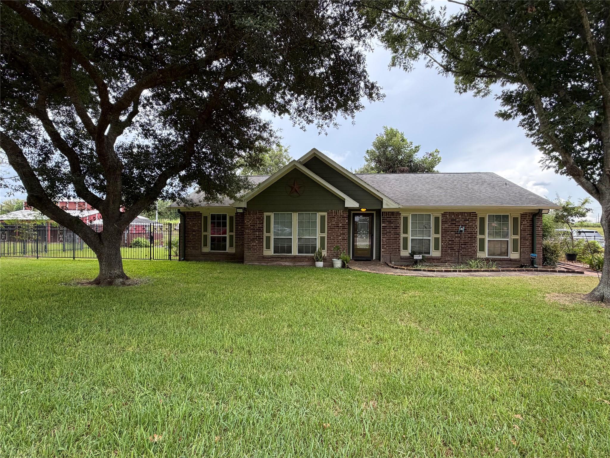Single-story brick house with a green gabled entry, large lawn, mature trees, and side fence.