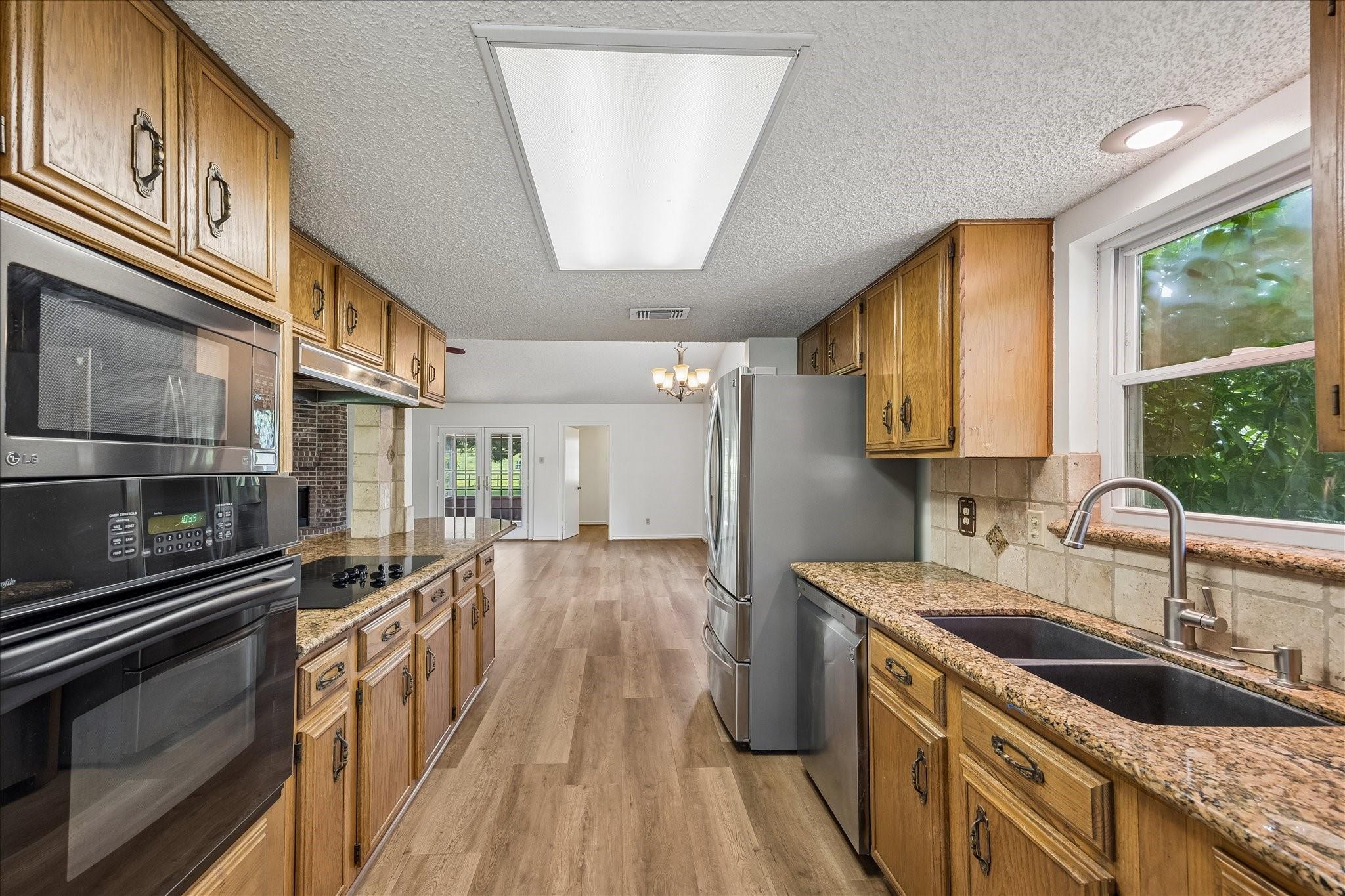 Galley kitchen with honey-colored cabinets, granite counters, stainless appliances, and a sink beneath a large window.