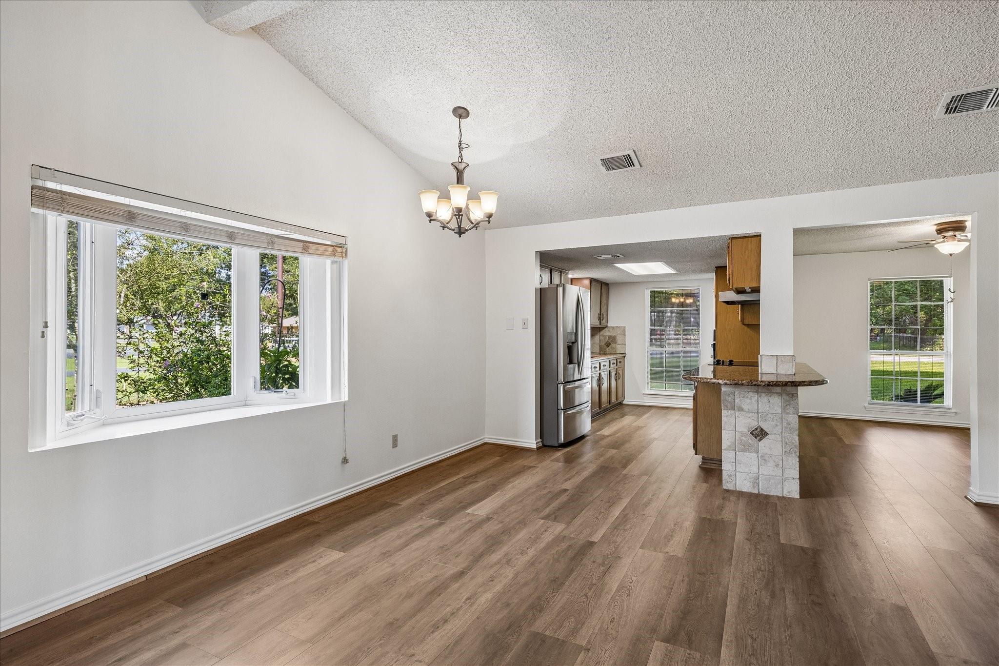 Empty dining area with wood-look floors, large windows, and adjoining kitchen with a tiled island.
