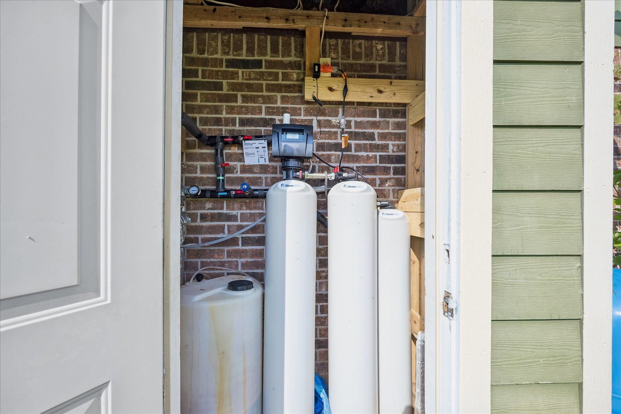 Outdoor water filtration system with several white tanks, plumbing, and controls inside a brick utility enclosure.