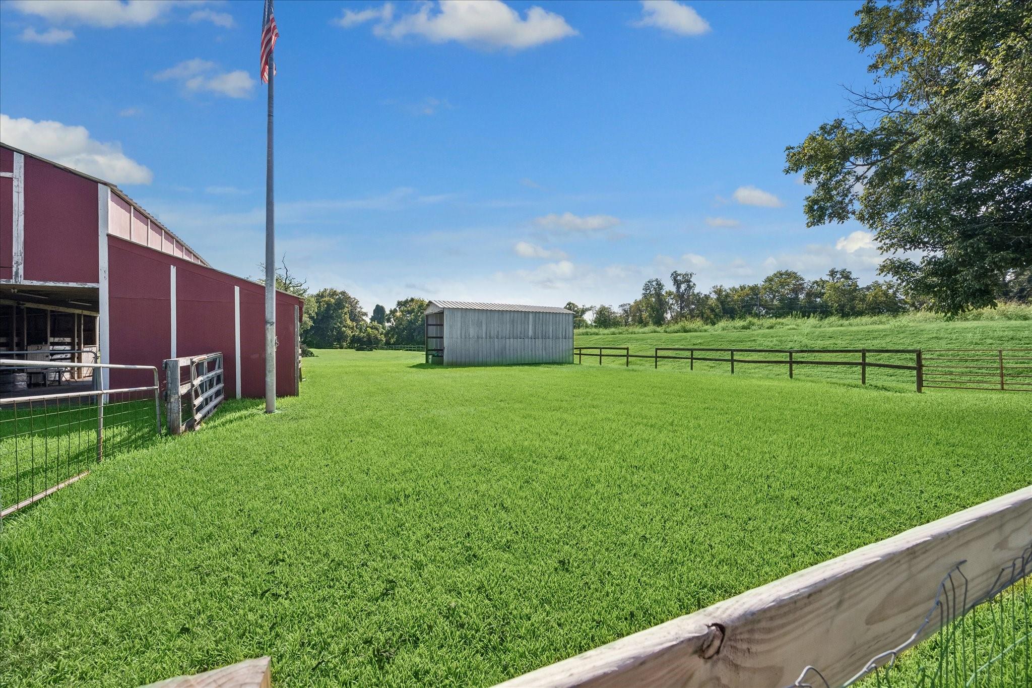 Fenced grassy paddock beside a red barn, with a gray shed and American flagpole.