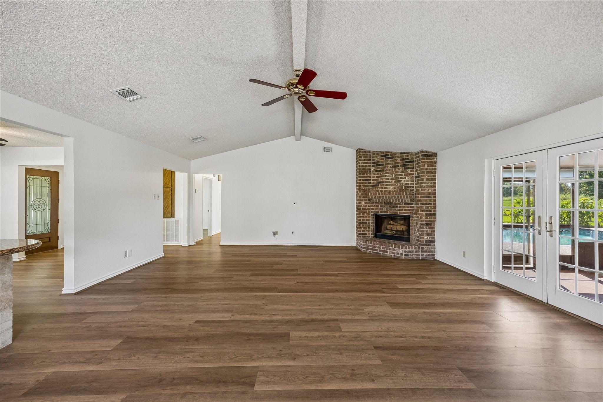 Living room with wood-look floors, a brick corner fireplace, and French doors overlooking a pool.