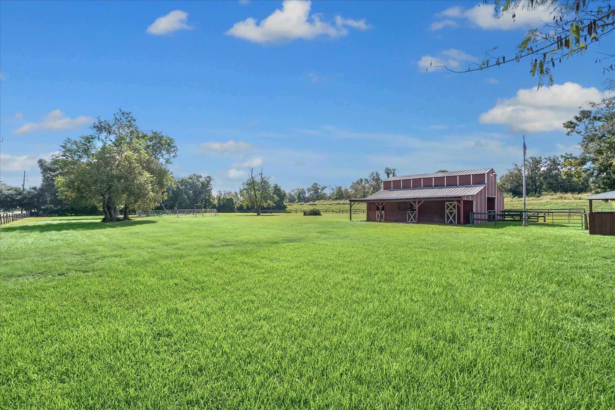 Red barn beside a wide green pasture with fenced enclosures and mature trees.