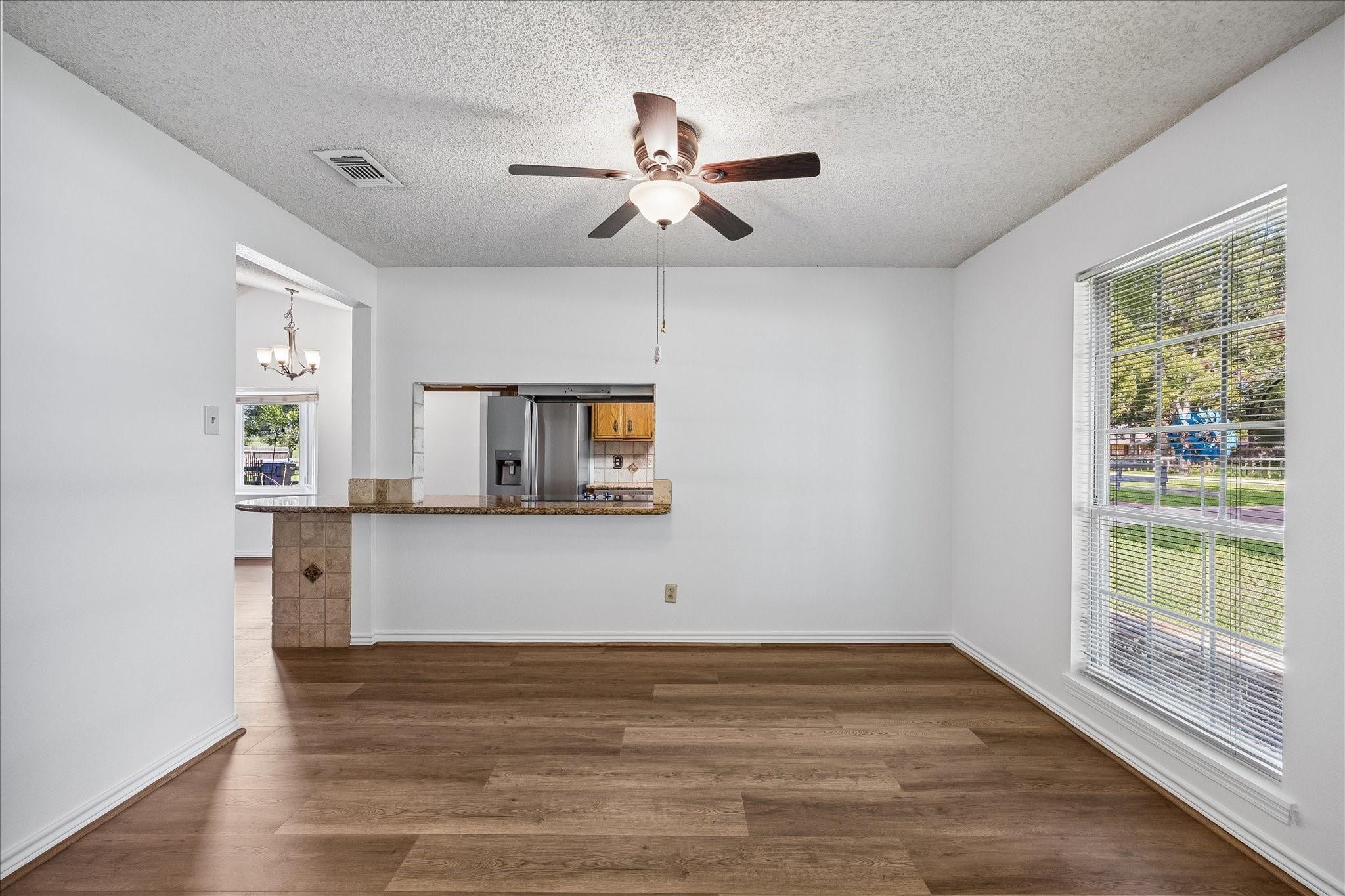 Vacant room with wood-look floors, ceiling fan, large window, and kitchen pass-through.