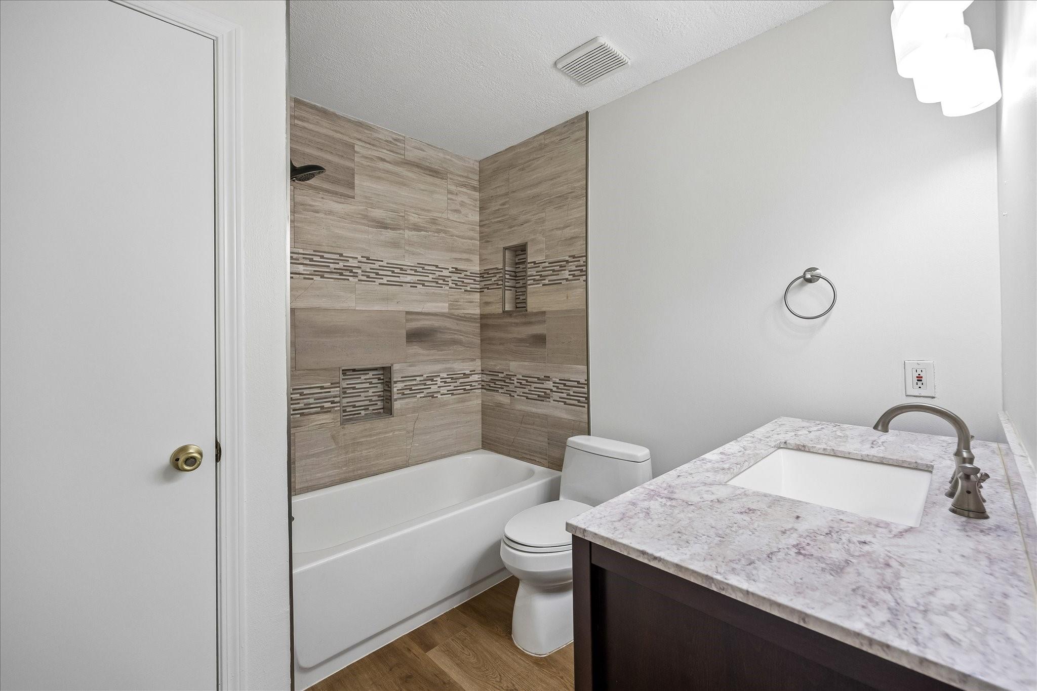 Bathroom with a tiled tub and shower, toilet, and dark vanity with a stone countertop.