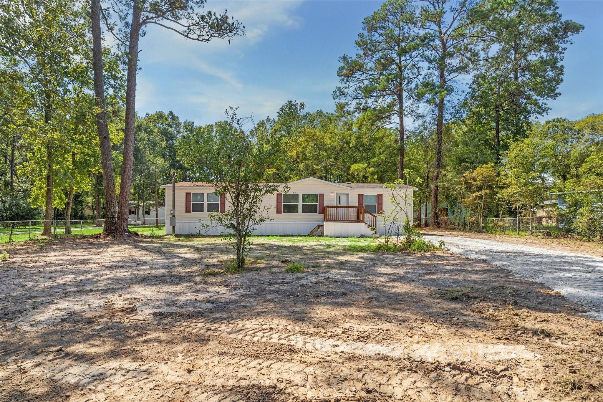 Manufactured home with red shutters and a small porch on a wooded lot beside a broad dirt driveway.
