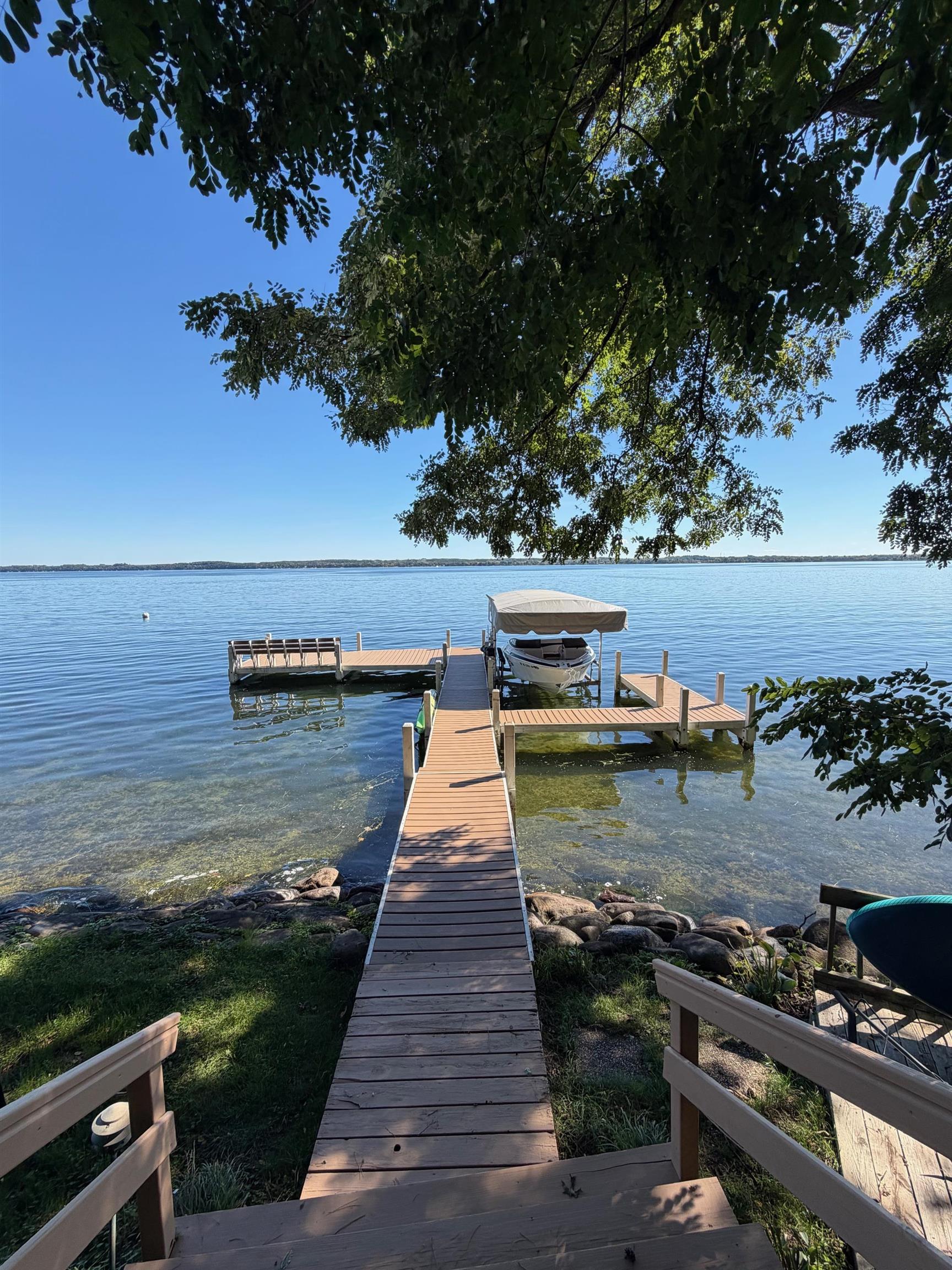 Lakeside dock with a covered boat beneath leafy trees on a clear day.
